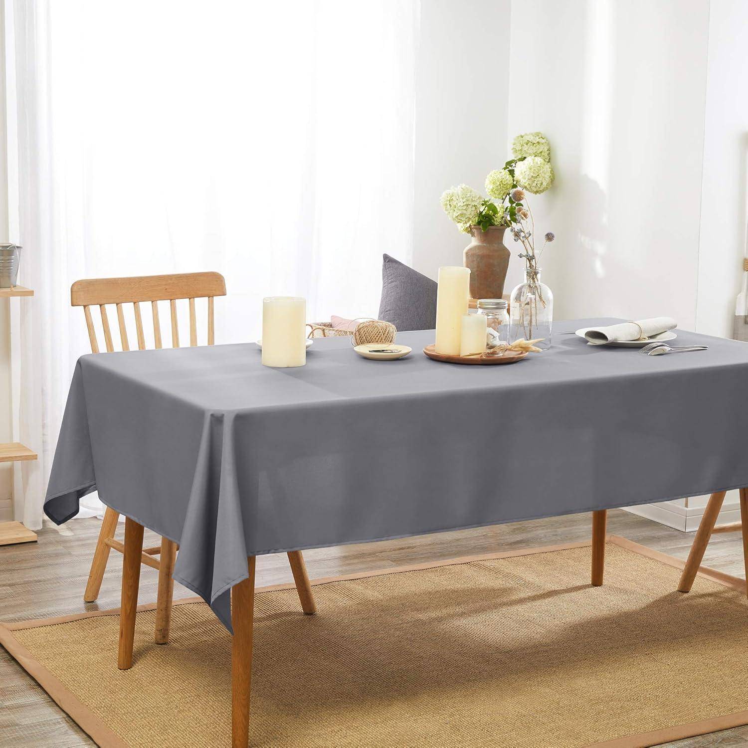 Esstisch mit einer grauen Tischdecke, zwei Kerzen, einer Vase mit Blumen, Tellern und einem Holzstuhl in einem hellen Raum mit Vorhängen.
