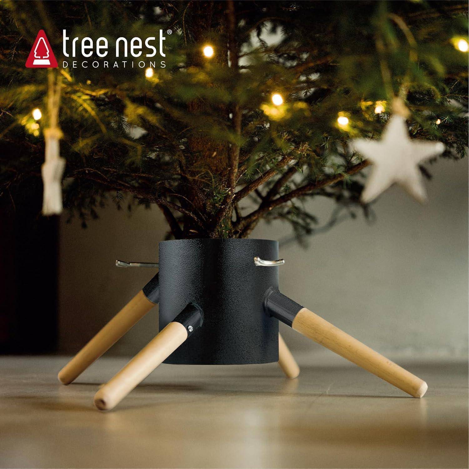 Ein geschmückter Weihnachtsbaum mit Lichtern steht in einem schwarzen Ständer mit Holzbeinen auf dem Boden. Das Logo "Tree Nest Decorations"" ist sichtbar.