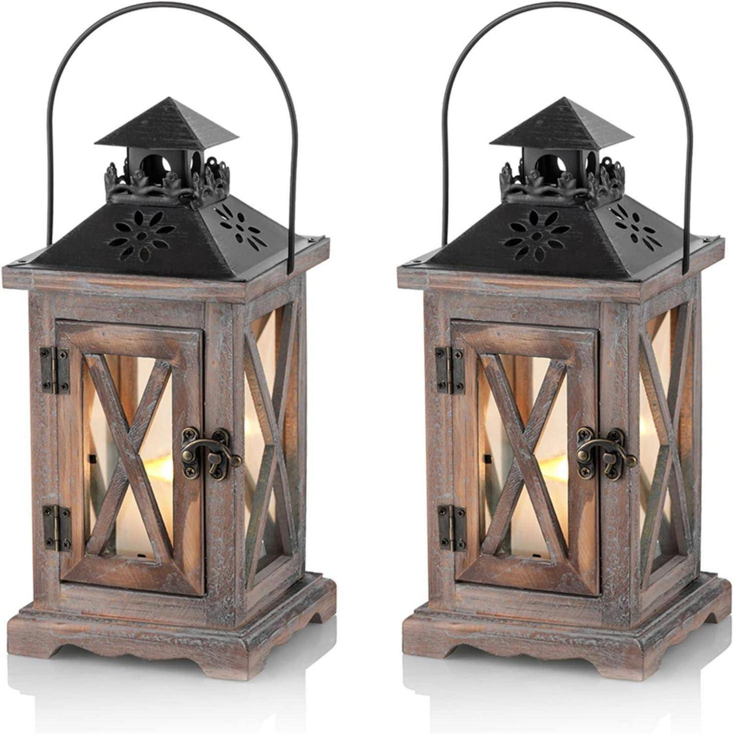 Zwei rustikale Holzlaternen mit schwarzen Metalldächern, ausgestattet mit Glaspaneelen und X-förmigen Holzverzierungen, in denen jeweils eine brennende Kerze steht.