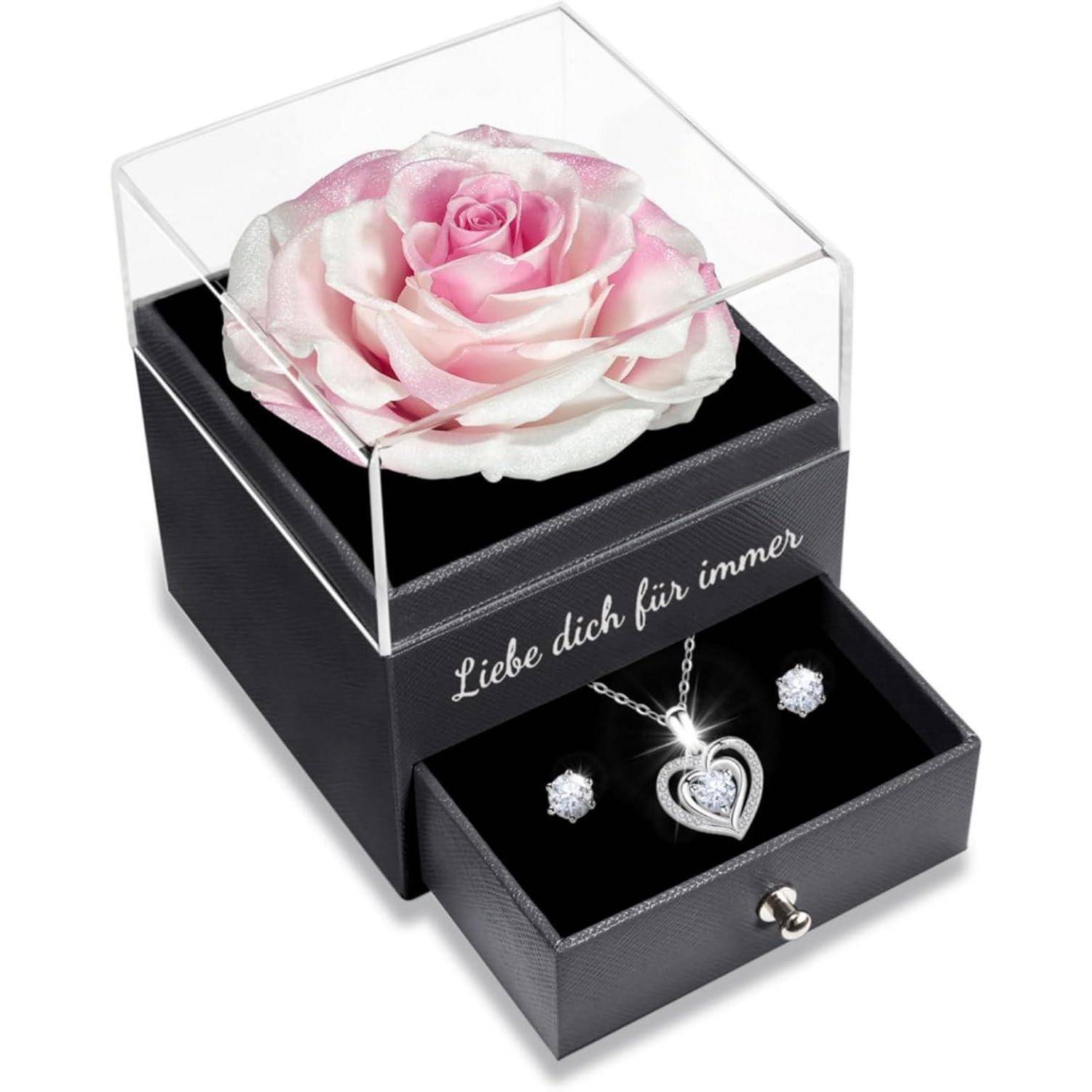 Eine rosa und weiße Rose in einer durchsichtigen Box auf einer schwarzen Schublade, die ein herzförmiges Anhänger und Ohrringe enthält. Die Box trägt die Aufschrift 'Liebe dich für immer'.