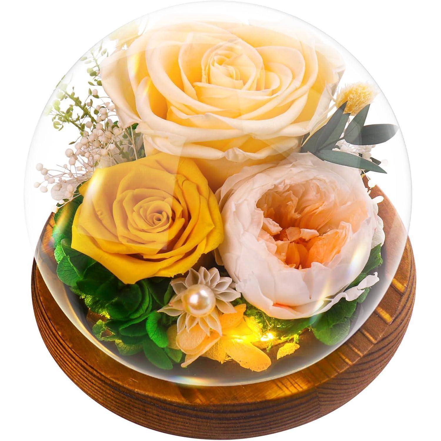Eine Glaskuppel umschließt konservierte gelbe und cremefarbene Rosen mit Grünzeug auf einer Holzbasis und schafft so eine elegante, zeitlose Blumenarrangement.