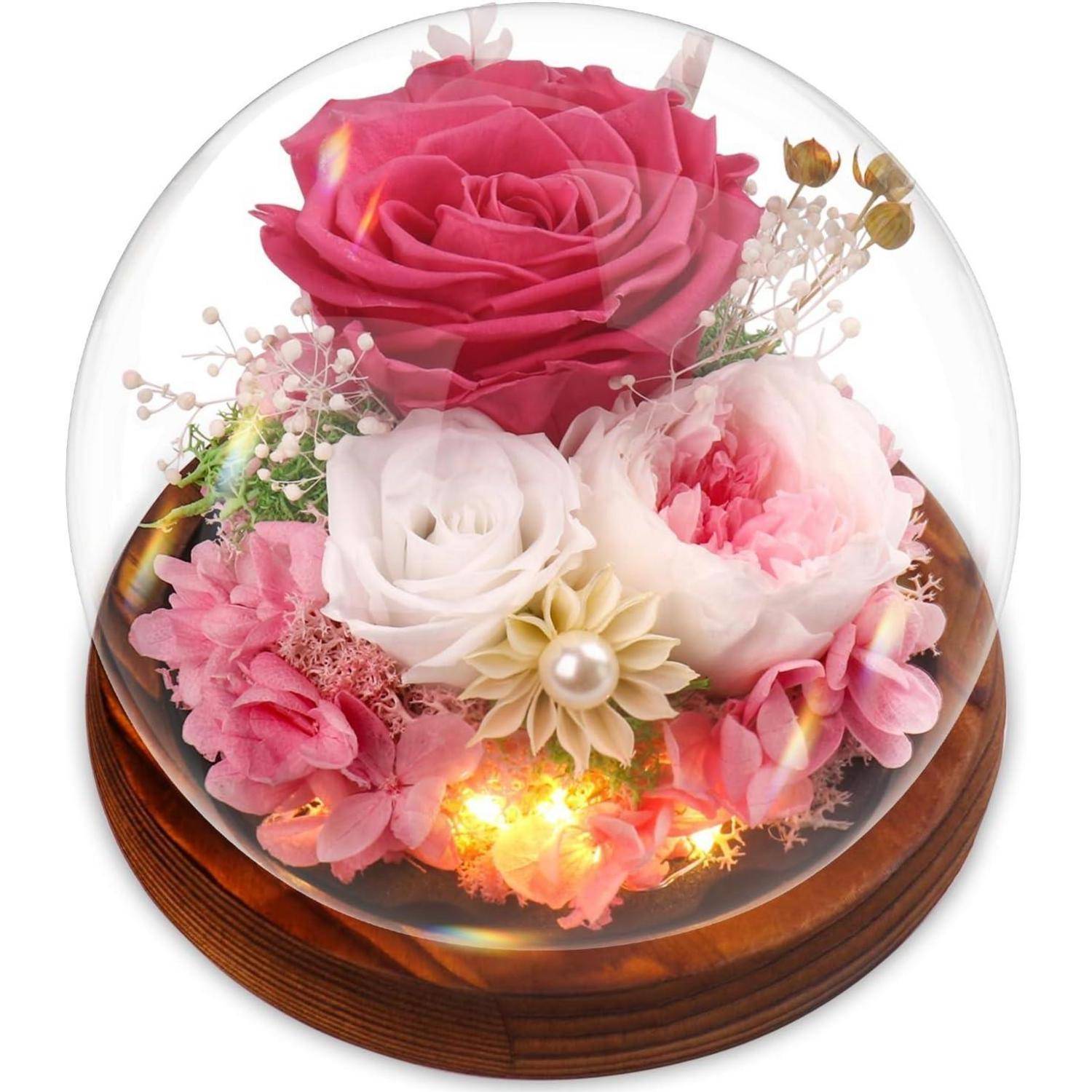 Eine Glasglocke umschließt eine Anordnung von rosa, weißen und roten Blumen mit kleinen dekorativen Lichtern auf einer Holzunterlage und unterstreicht damit Eleganz.