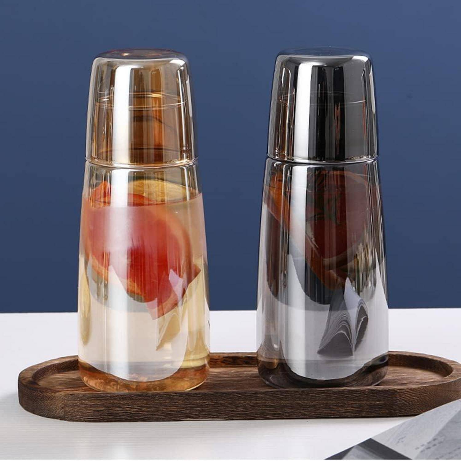 Zwei Glasbecher mit Doppelwänden, auf einem Holztablett umgedreht aufgestellt, die ein abstraktes Design reflektieren; vor einem blauen Hintergrund.