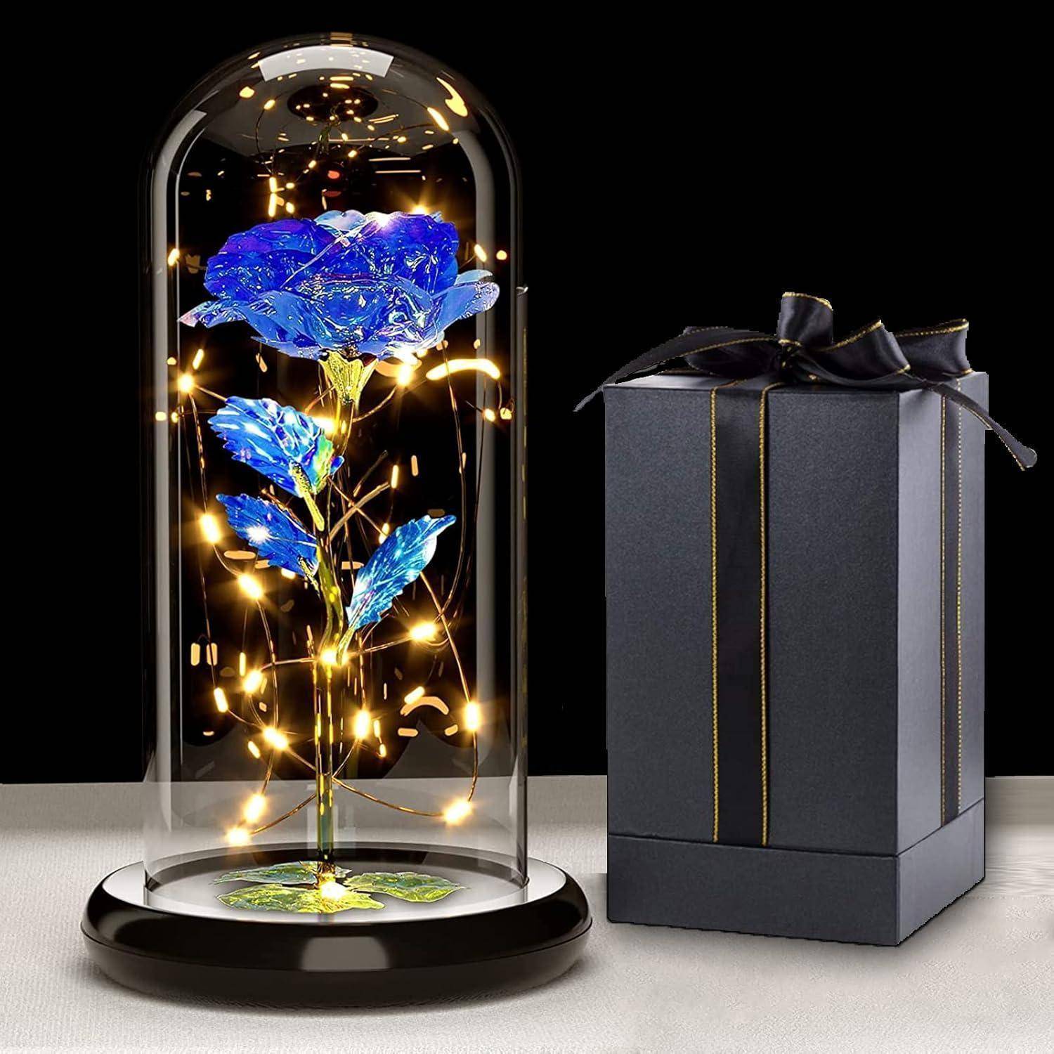 Eine Glaskuppel umschließt eine leuchtende blaue Rose, ausgestattet mit LED-Lichtern. Daneben sitzt eine schwarze Geschenkbox mit einem Band auf einem Tisch.