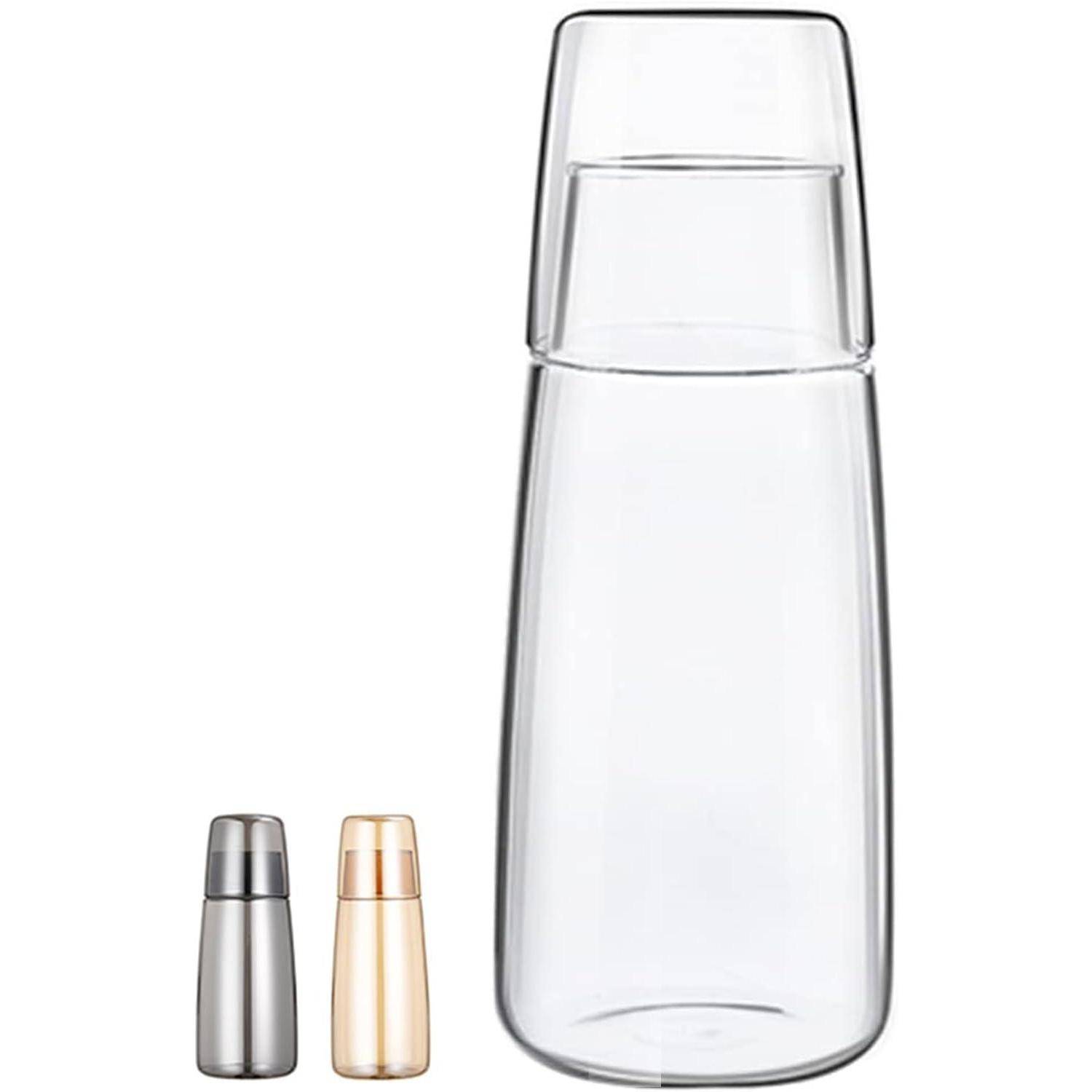 Eine hohe, transparente Glasflasche mit einer passenden, darauf umgedrehten Glasschale. Zwei kleinere Flaschen, eine silberne und eine goldene, befinden sich daneben.