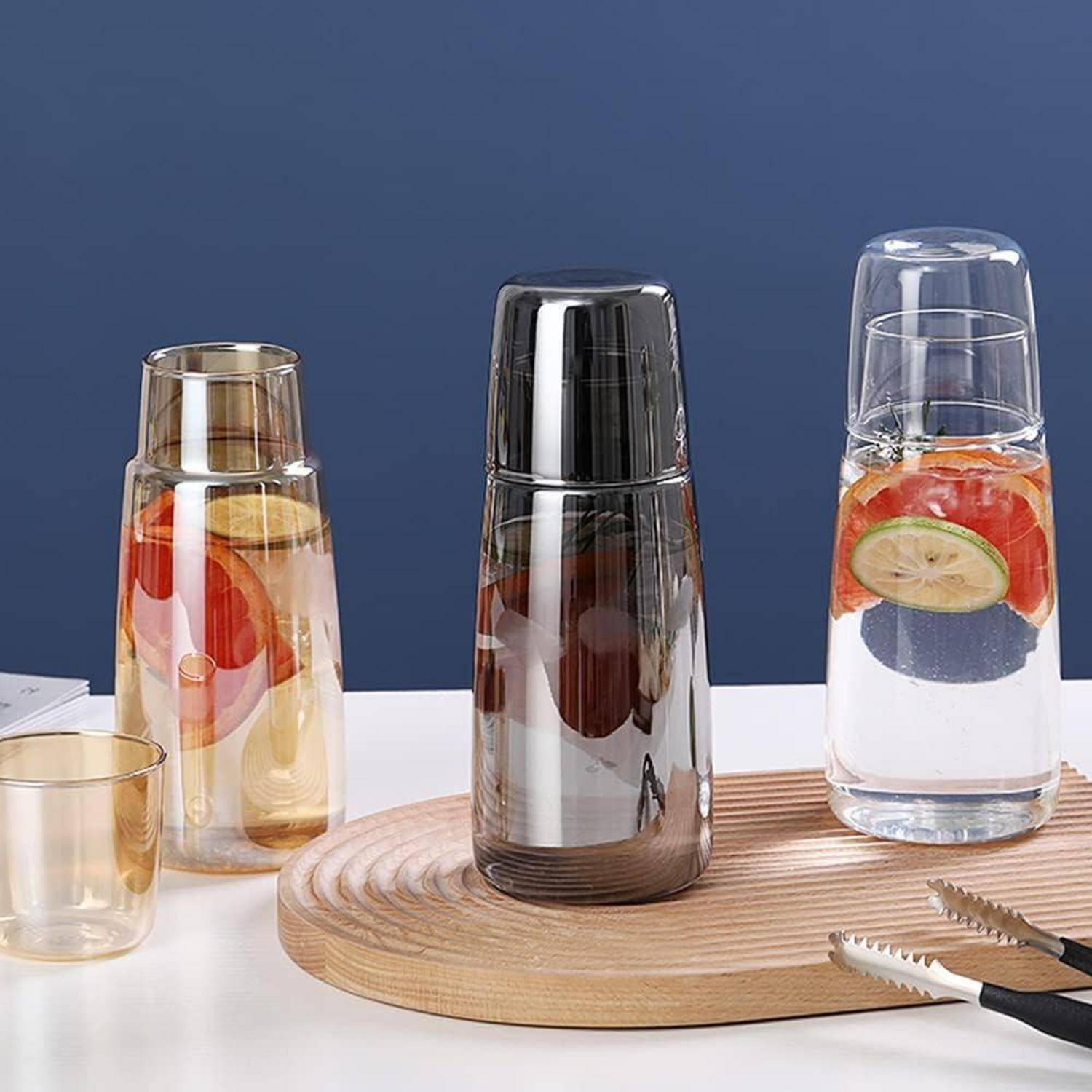Drei Glasflaschen, jede mit einem Deckel und gefüllt mit Wasser und Fruchtscheiben, stehen auf einem Holztablett mit Küchenzange.