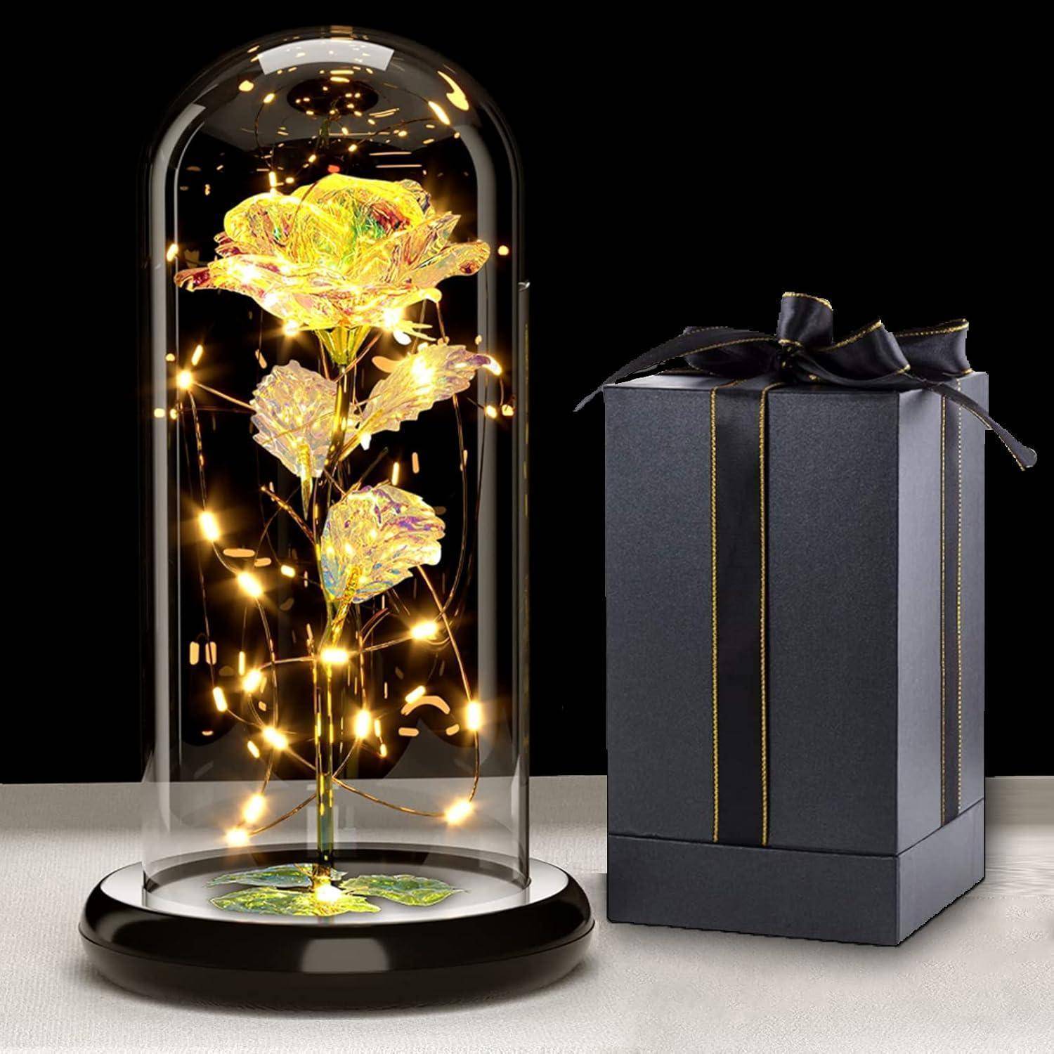 Eine Glaskuppel umschließt eine leuchtende, goldene Rose mit Feenlichtern. Eine schwarze Geschenkbox mit einer Schleife sitzt daneben.