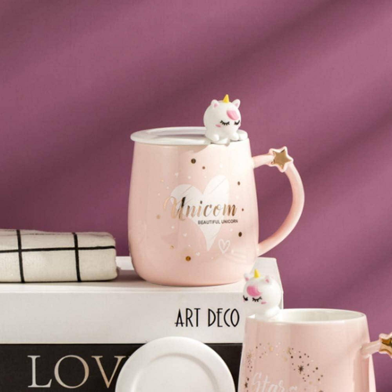 Eine rosa Tasse mit Einhorn-Motiv, auf der der Text "Beautiful Unicorn"" steht, neben einem Handtuch und einer weiteren ähnlichen Tasse vor einem malvenfarbenen Hintergrund.