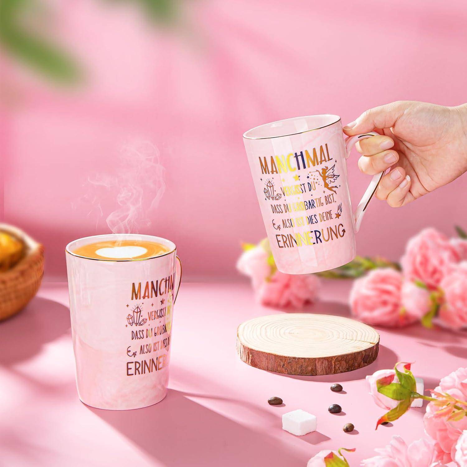Eine Hand hält eine rosa Tasse mit Text. Eine weitere Tasse steht daneben, aus der Dampf aufsteigt. Der Hintergrund zeigt rosa Blumen und Holzuntersetzer.