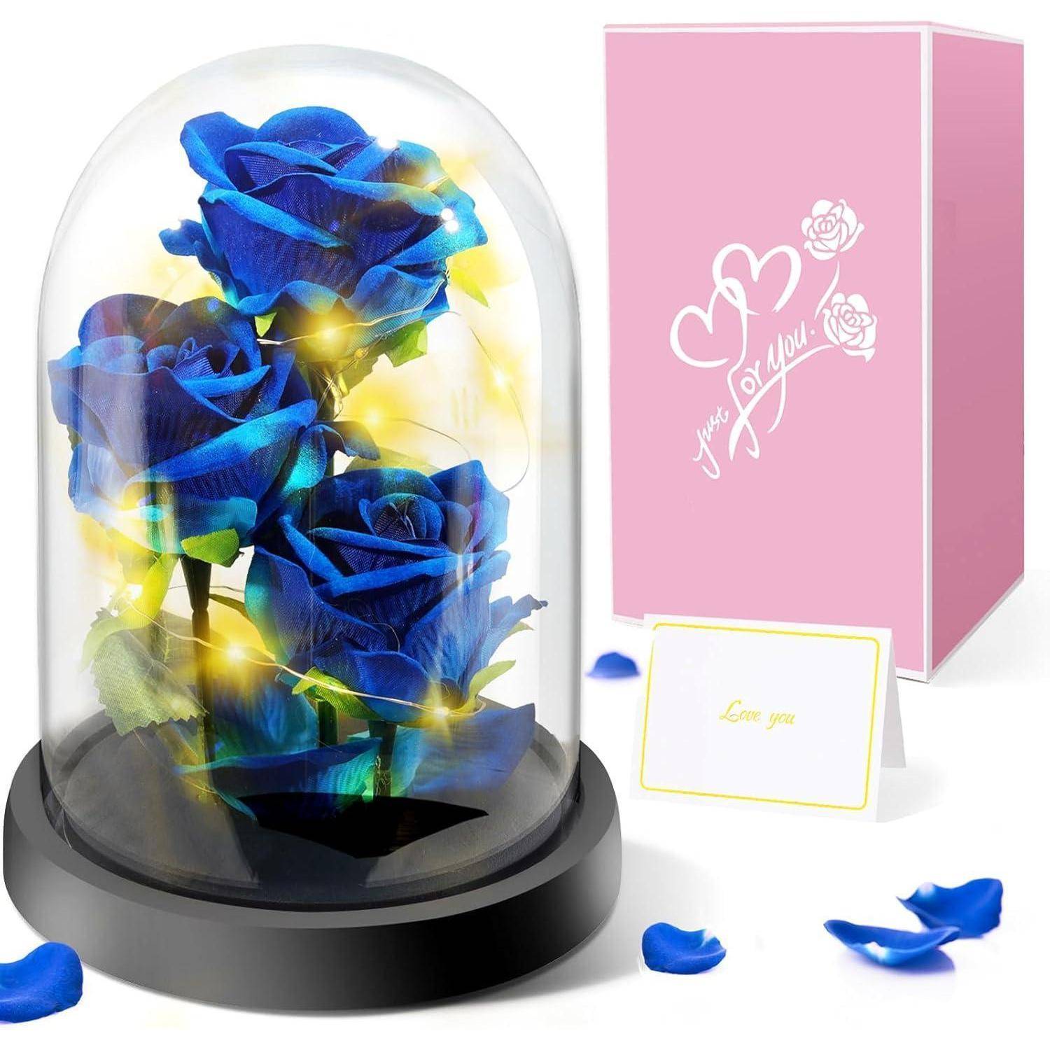Eine Glaskuppel umschließt beleuchtete blaue Rosen auf einem schwarzen Sockel. Eine rosa Schachtel mit der Aufschrift ‚Nur für dich!' und eine Karte mit der Aufschrift ‚Ich liebe dich' befinden sich in der Nähe.