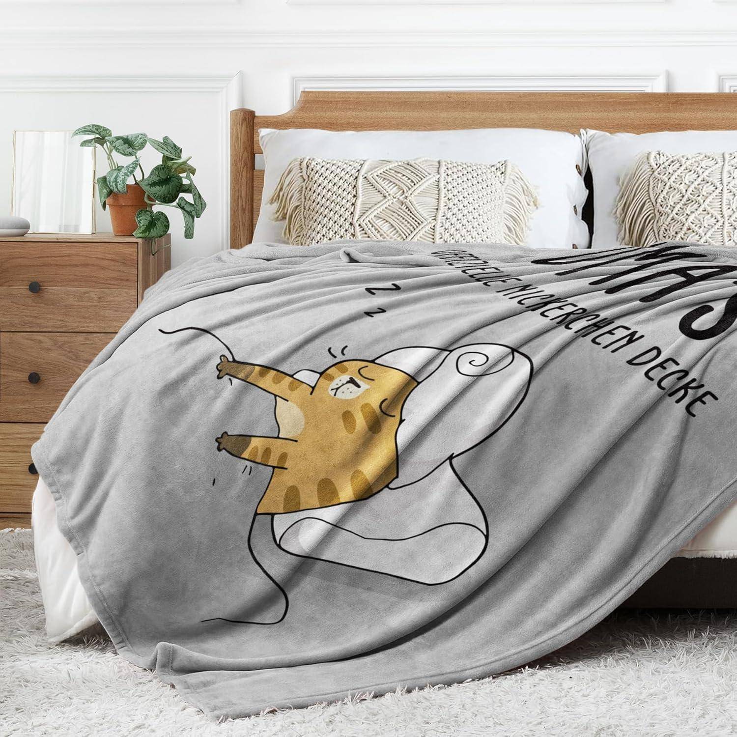 Eine gemütliche Schlafzimmer-Szene mit einem Bett, das mit einer dicken grauen Decke ausgestattet ist, die eine Cartoon-Katze-Illustration und Text auf Deutsch zeigt.