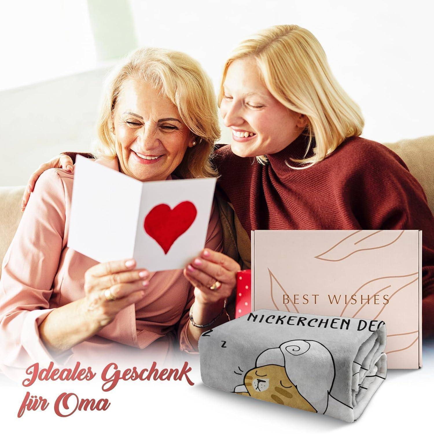 Eine ältere Frau und eine jüngere Frau sitzen zusammen, lächeln und schauen auf eine Karte mit einem roten Herz. Eine Geschenkdecke mit der Aufschrift 