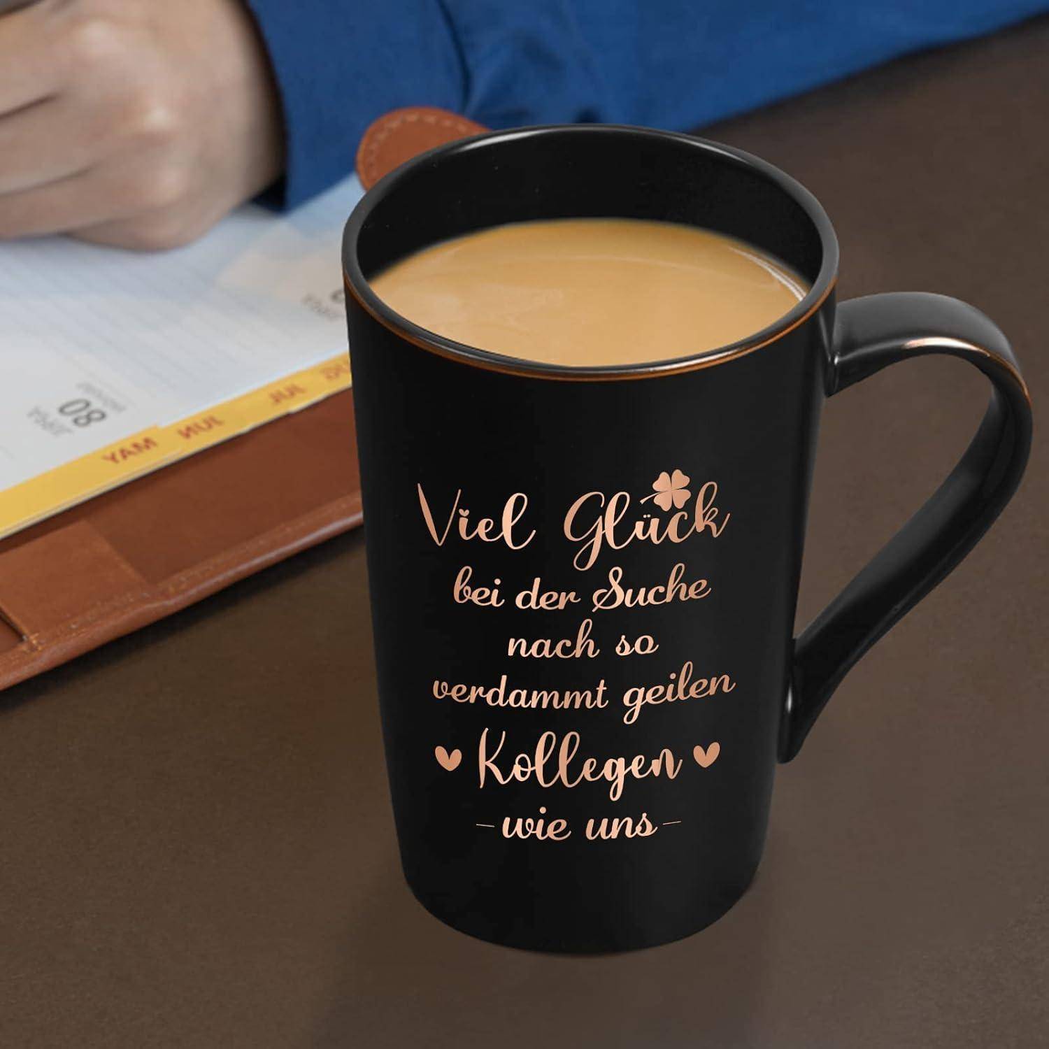 Eine schwarze Tasse mit deutschem Text 'Viel Glück bei der Suche nach so verdammt geilen Kollegen wie uns', gefüllt mit Kaffee. Daneben schreibt eine Hand in einem Planer.