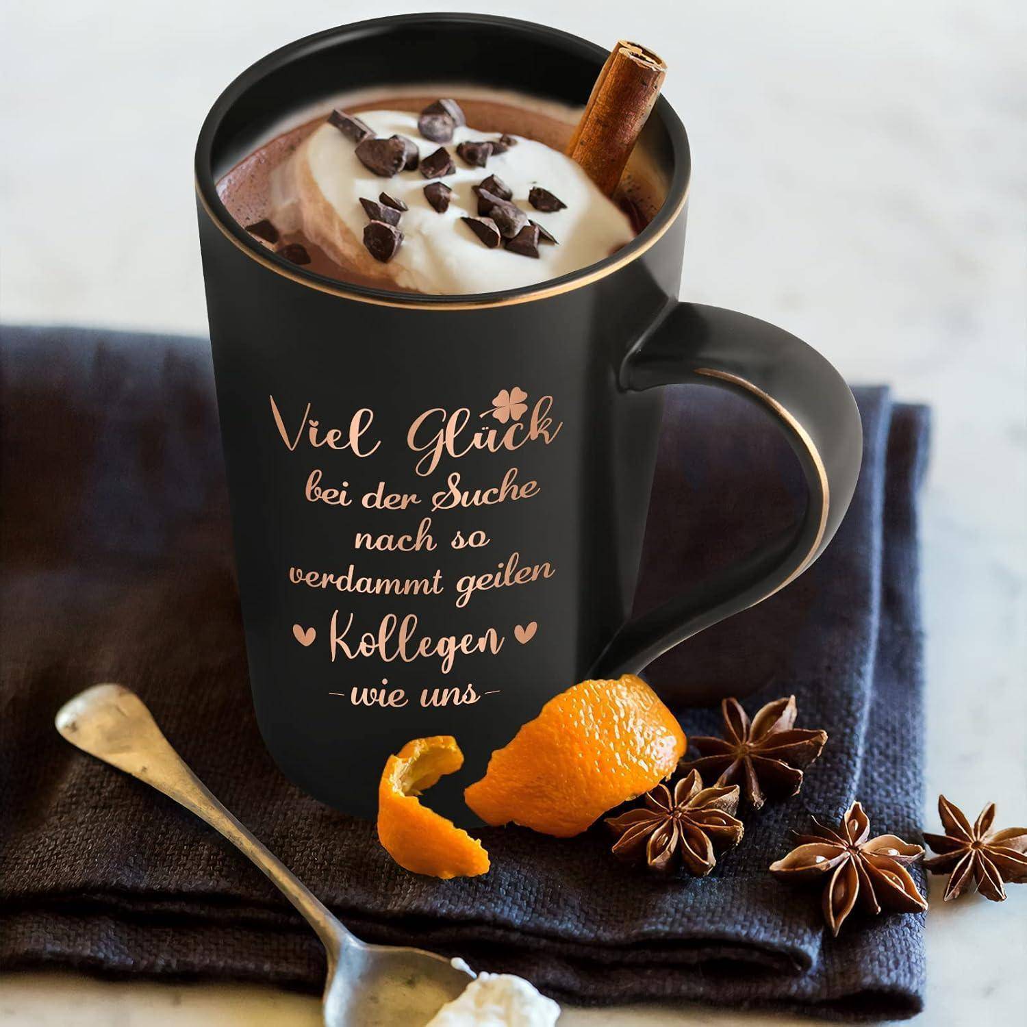 Eine schwarze Kaffeetasse mit heißer Schokolade, gekrönt von Schlagsahne, Schokoladenspänen und einem Zimtstange. Text auf der Tasse: 'Viel Glück bei der Suche nach so verdammt geilen Kollegen wie uns.' Eine Orangenschale und ein Sternanis neben der Tasse.
