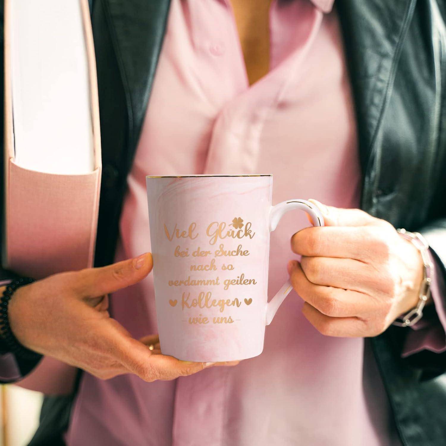 Eine Person in einem pinken Hemd und einer schwarzen Jacke hält eine Tasse mit deutschem Text und trägt eine pinke Mappe, was eine professionelle Umgebung vermittelt.