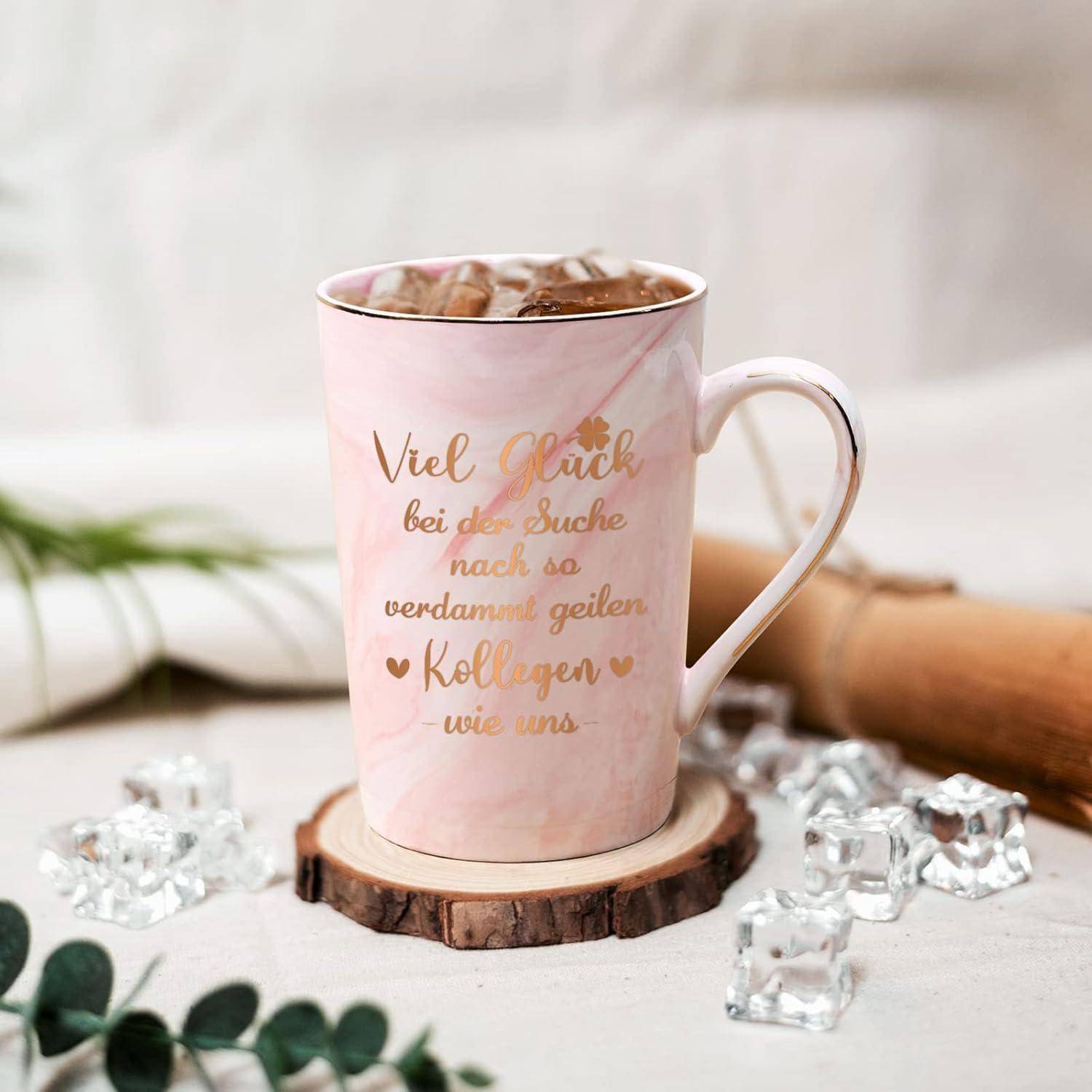 Eine pinke Tasse mit goldener Aufschrift, auf der steht 'Viel Glück bei der Suche nach so verdammt geilen Kollegen wie uns', auf einem Holzuntersetzer, umgeben von dezenter Dekoration.