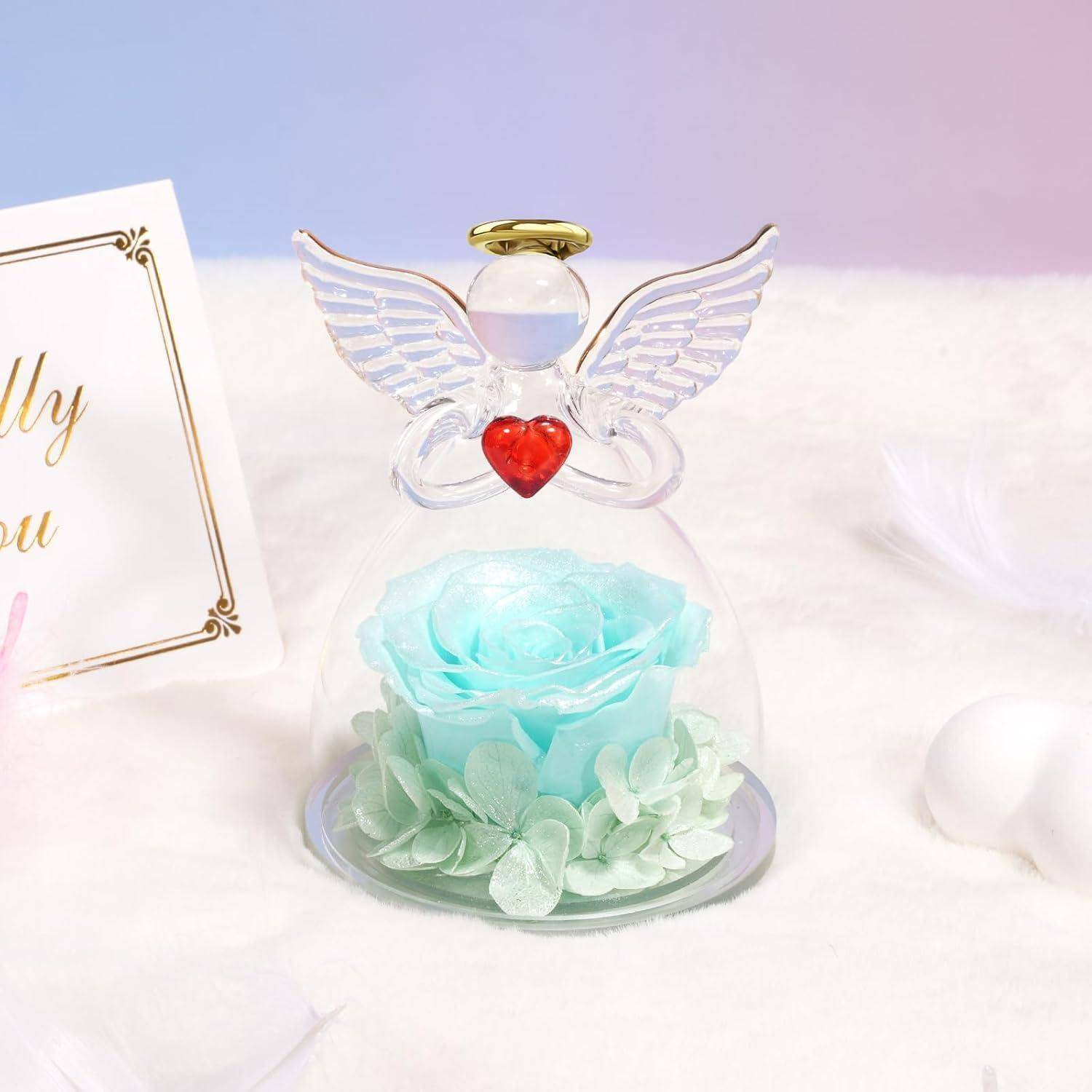 Eine Glasfigur eines Engels mit einem roten Herzen und einer leuchtenden türkisfarbenen Rose steht neben einer Karte mit Schreibschrifttext auf einem weichen, pastellfarbenen Hintergrund.