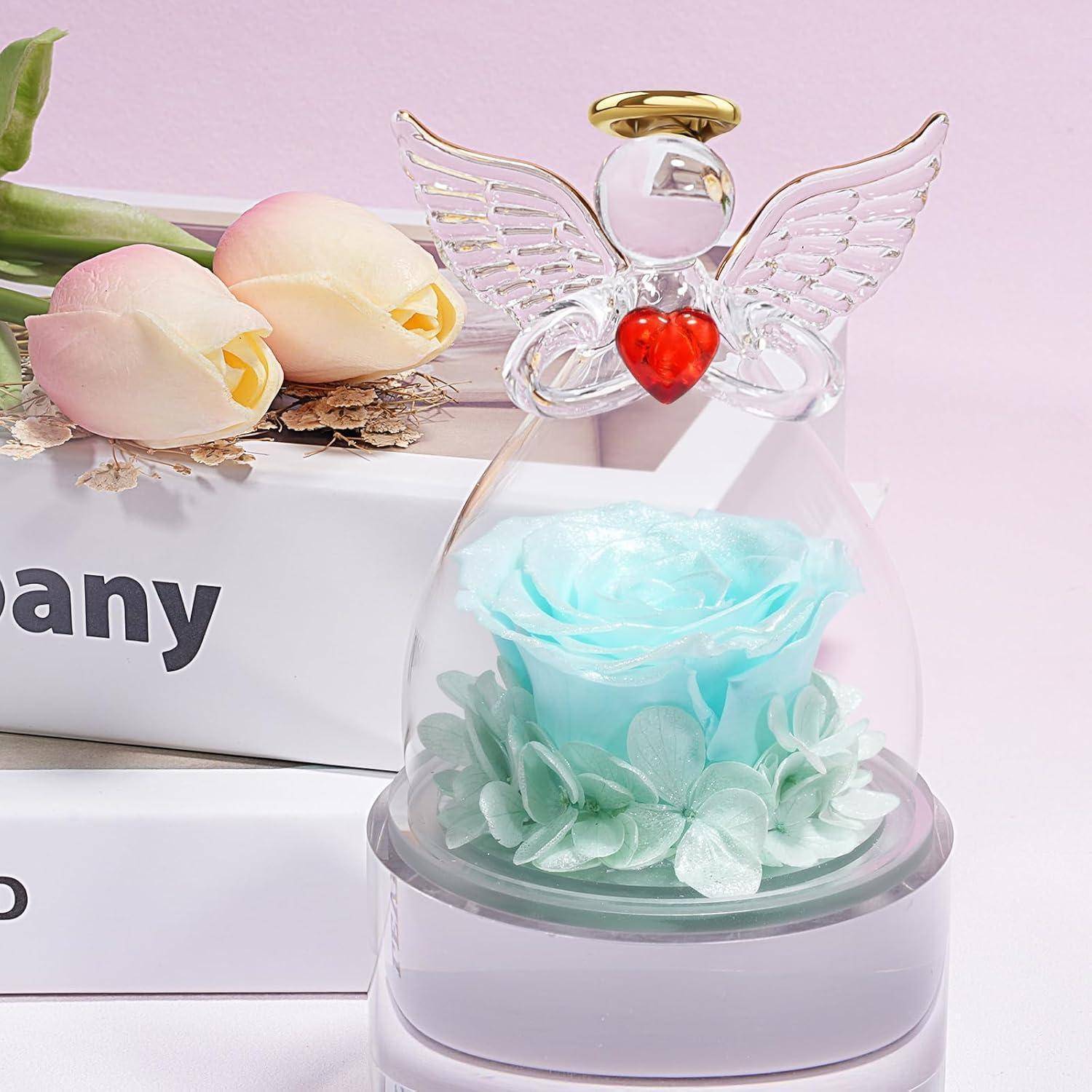 Eine dekorative Glasengelfigur mit Flügeln und einem roten Herzen umschließt eine blaue Rose, die sich vor rosa Tulpen und Büchern befindet.