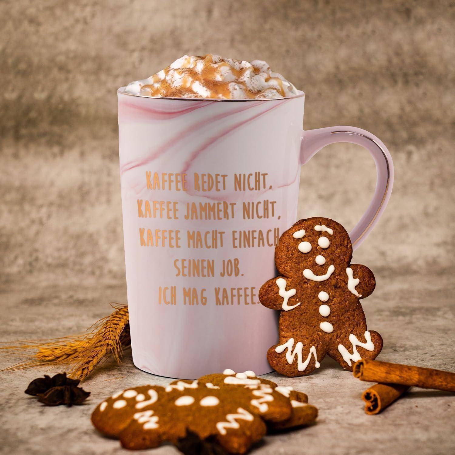 Eine rosa Marmortasse mit Schlagsahne und einem deutschen Zitat über Kaffee. Ein Lebkuchen, Weizen und Zimtstangen liegen daneben.