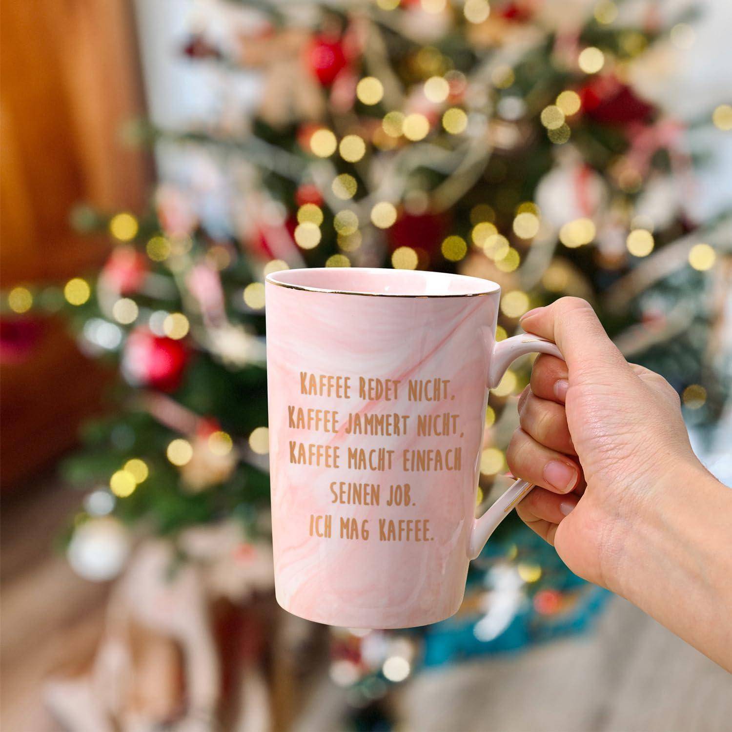 Eine Hand hält eine weiße Kaffeetasse mit goldener Schrift. Im Hintergrund ist ein geschmückter Weihnachtsbaum leicht unscharf.