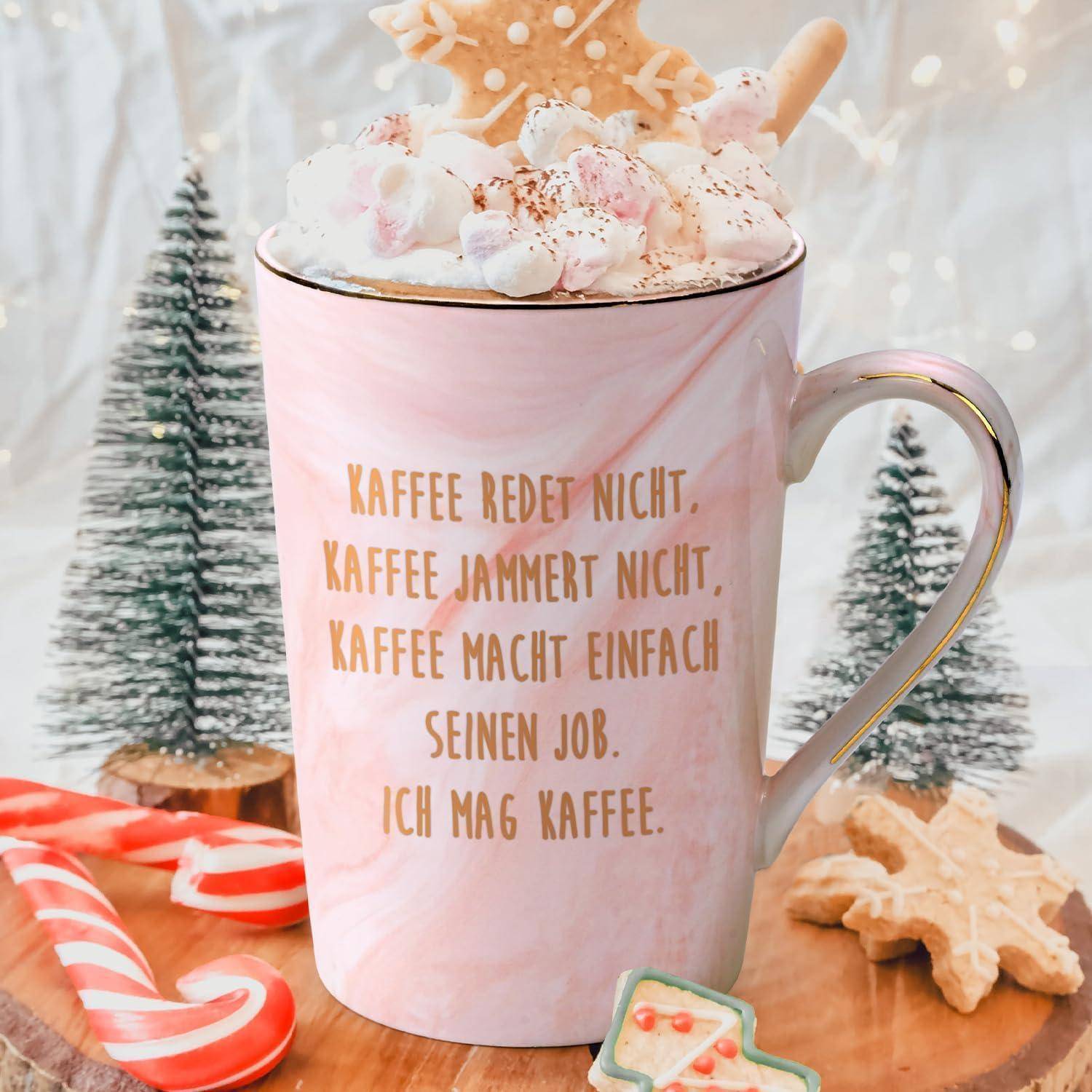 Eine rosa Tasse gefüllt mit Schlagsahne und Marshmallows, umgeben von festlichen Dekorationen und kleinen gefrosteten Bäumen, mit Text auf Deutsch über Kaffee.