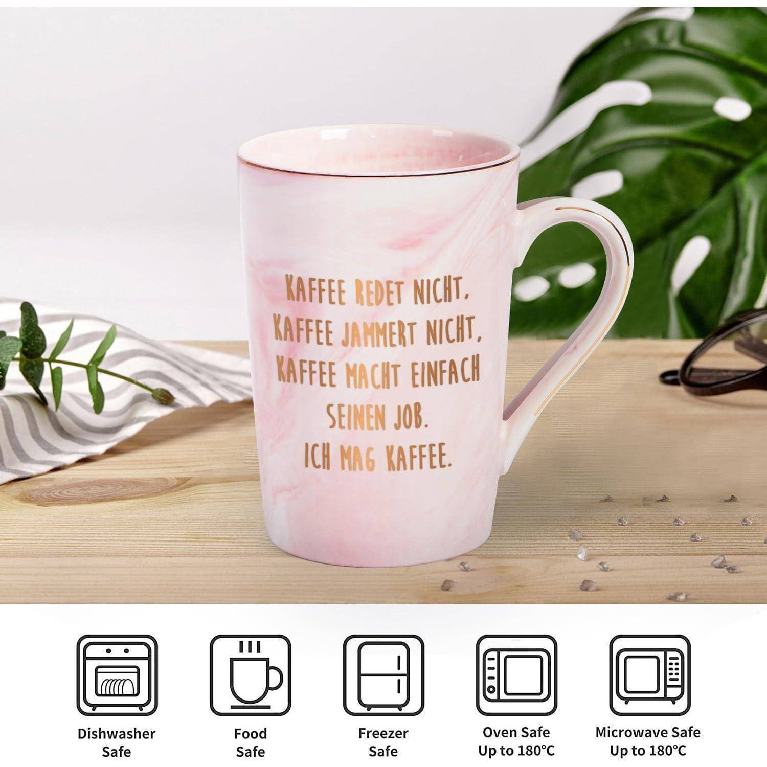 Eine rosa Keramiktasse mit deutschem Text über Kaffee, die auf einem Holztisch steht. Darunter sind Symbole für Spülmaschinen-, Gefrier-, Backofen- und Mikrowellensicherheit bis zu 180°C.