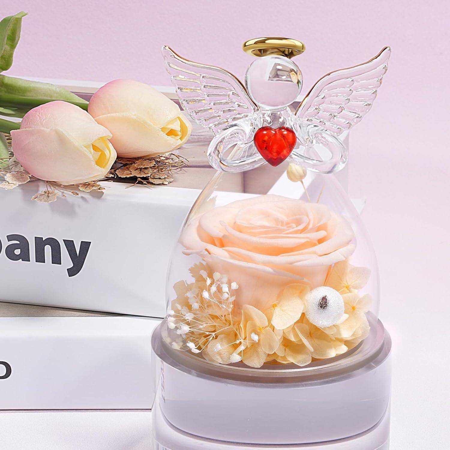 Gläserne Engelfigur mit Herz und Flügeln umschließt eine rosa Rose auf einem Stapel Bücher neben zwei Tulpen, vor einem weichen rosa Hintergrund.