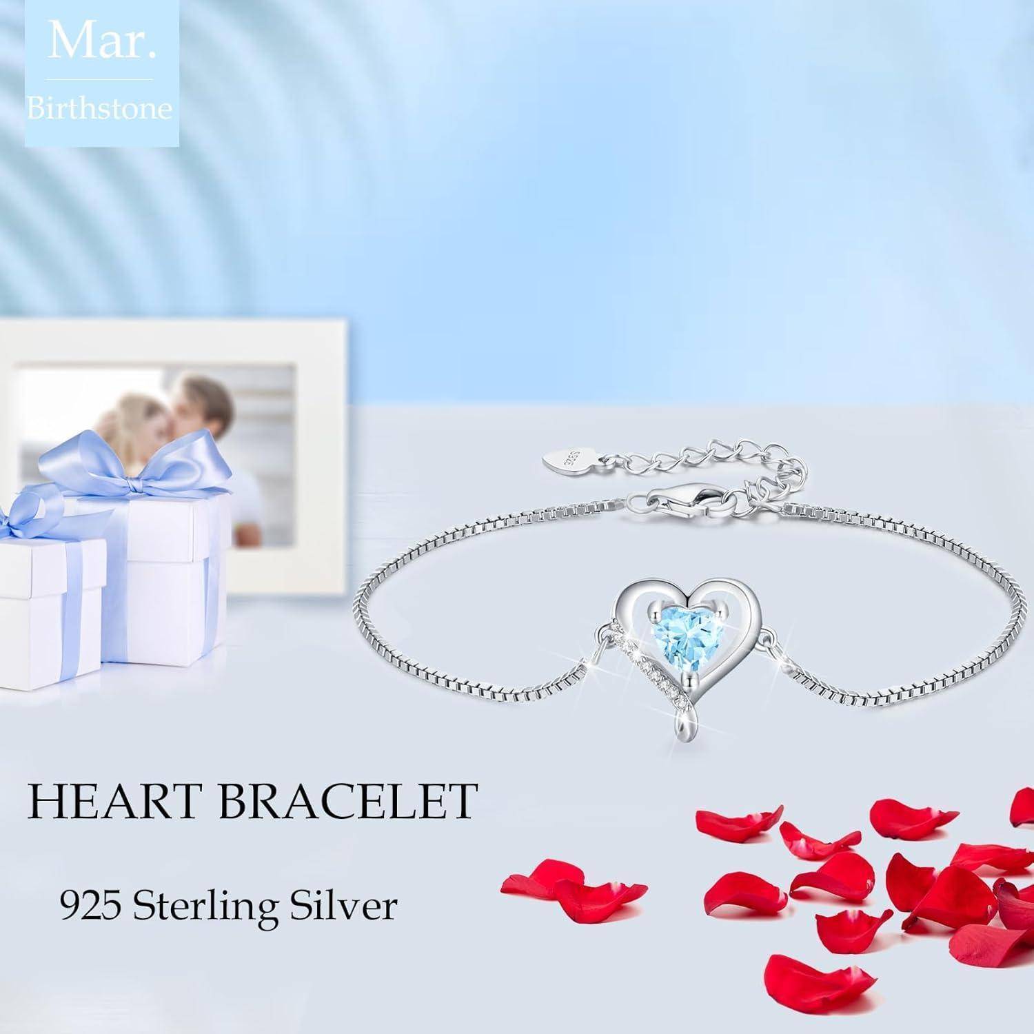 Ein Silberarmband in Herzform mit einem hellblauen Edelstein in der Mitte, positioniert neben einer kleinen weißen Geschenkbox mit einer blauen Schleife und roten Rosenblütenblättern.