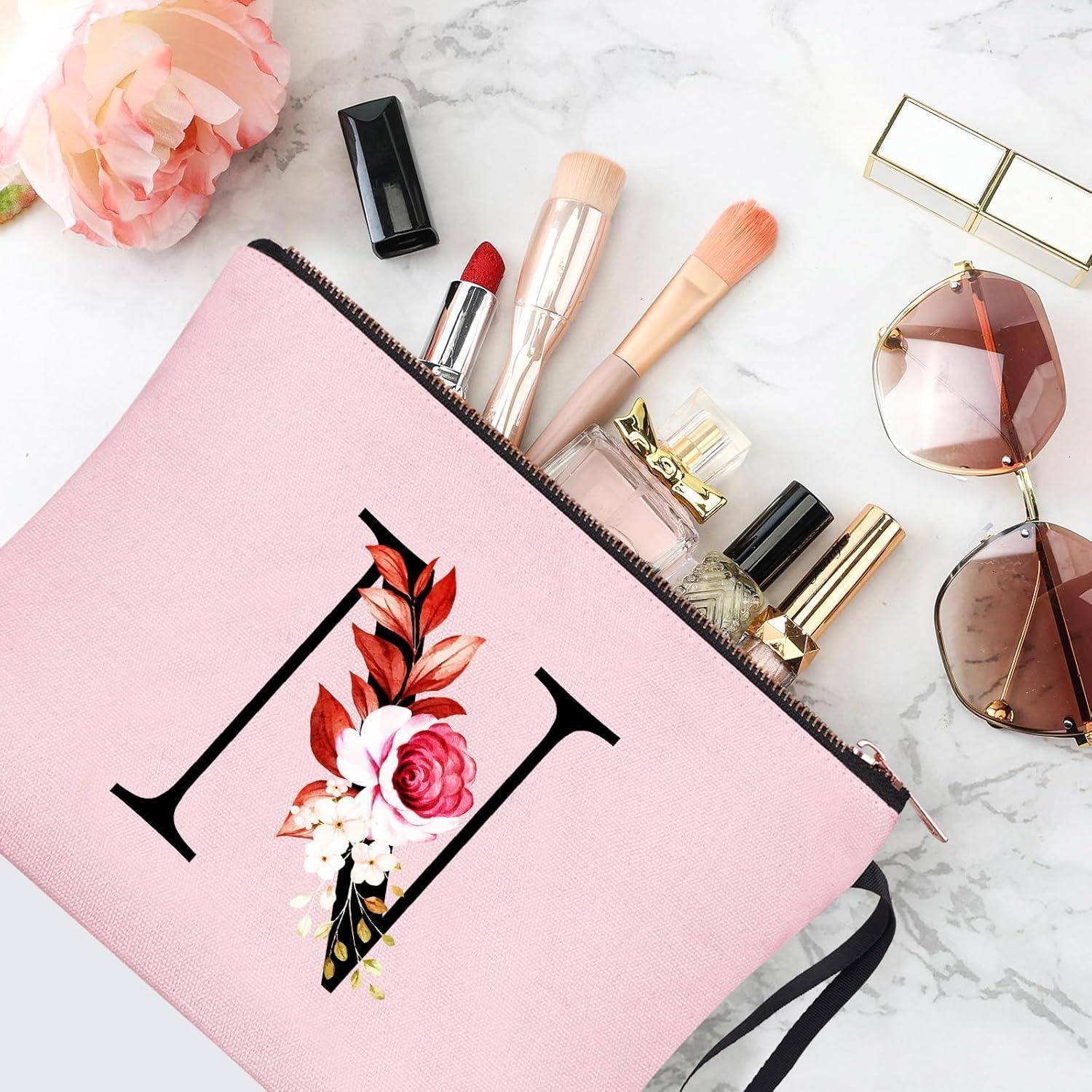 Eine rosa Kosmetiktasche mit einem floralen 'H' Monogramm ist geöffnet und zeigt Kosmetikprodukte wie Lippenstift, Mascara und Pinsel, umgeben von Sonnenbrille und einer Rose.