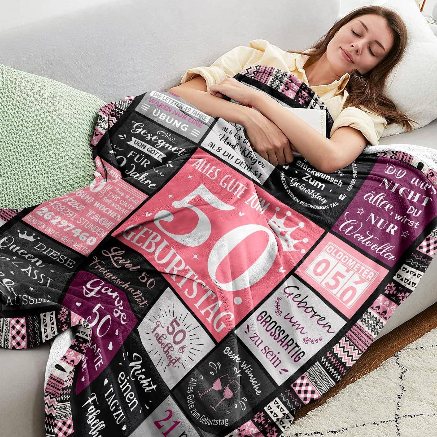 Eine Frau schläft auf einem Sofa, bedeckt mit einer Steppdecke, die deutsche Geburtstagswünsche und die Zahl '50' trägt.