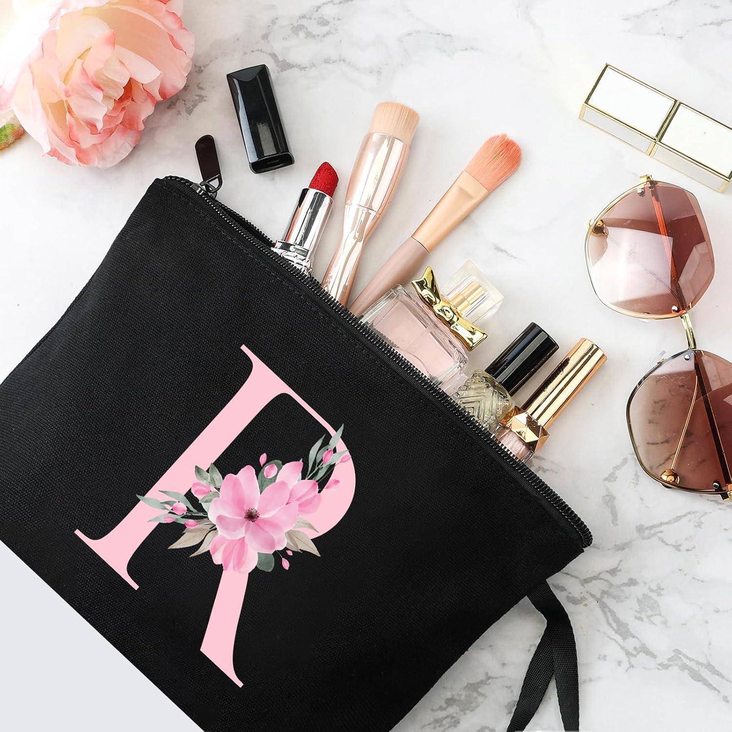 Eine schwarze Make-up-Tasche mit einem pinken floralen 'R'-Monogramm, umgeben von Kosmetikprodukten, Sonnenbrille und einer pinken Blume auf einer Marmoroberfläche.