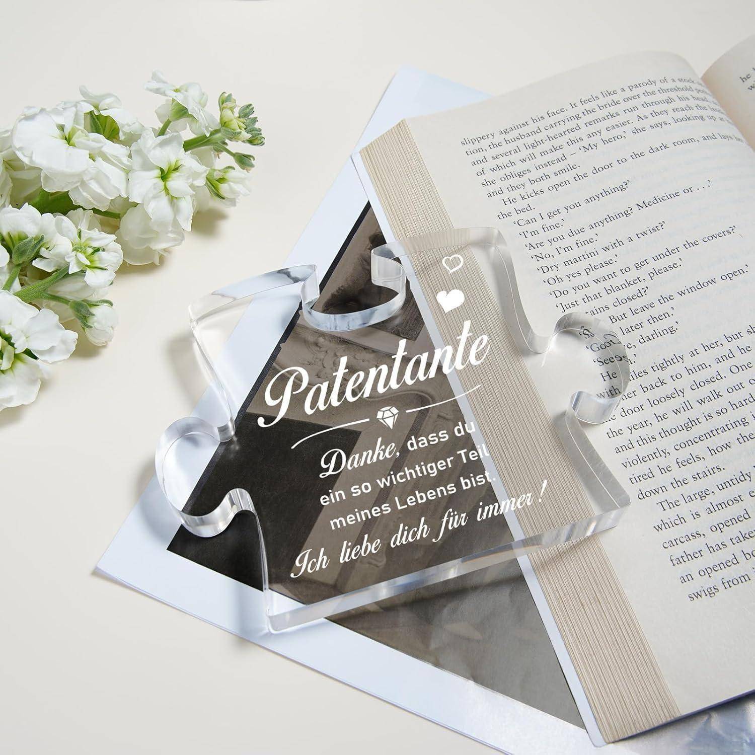 Ein transparentes Puzzleteil mit deutschem Text liegt auf einem aufgeschlagenen Buch. Daneben sind kleine weiße Blumen dekorativ angeordnet.