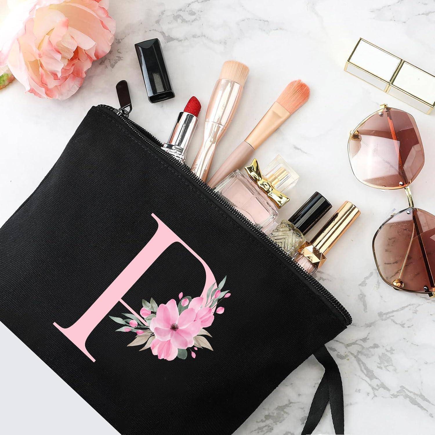 Kosmetiktasche mit dem Buchstaben 'L' und floralem Design, die Lippenstift, Pinsel, Parfüm und Mascara enthält. Sonnenbrille und eine Blume in der Nähe.