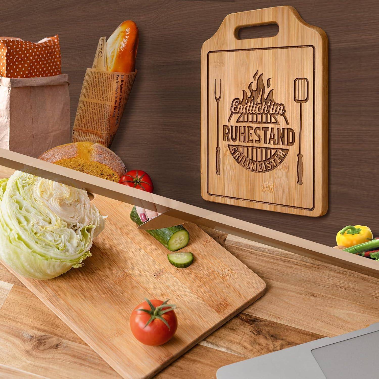 Ein Schneidebrett mit dekorativem Text und Küchenutensilien wird neben einem Laib Brot, Salat, Gurkenscheiben und Tomaten präsentiert.