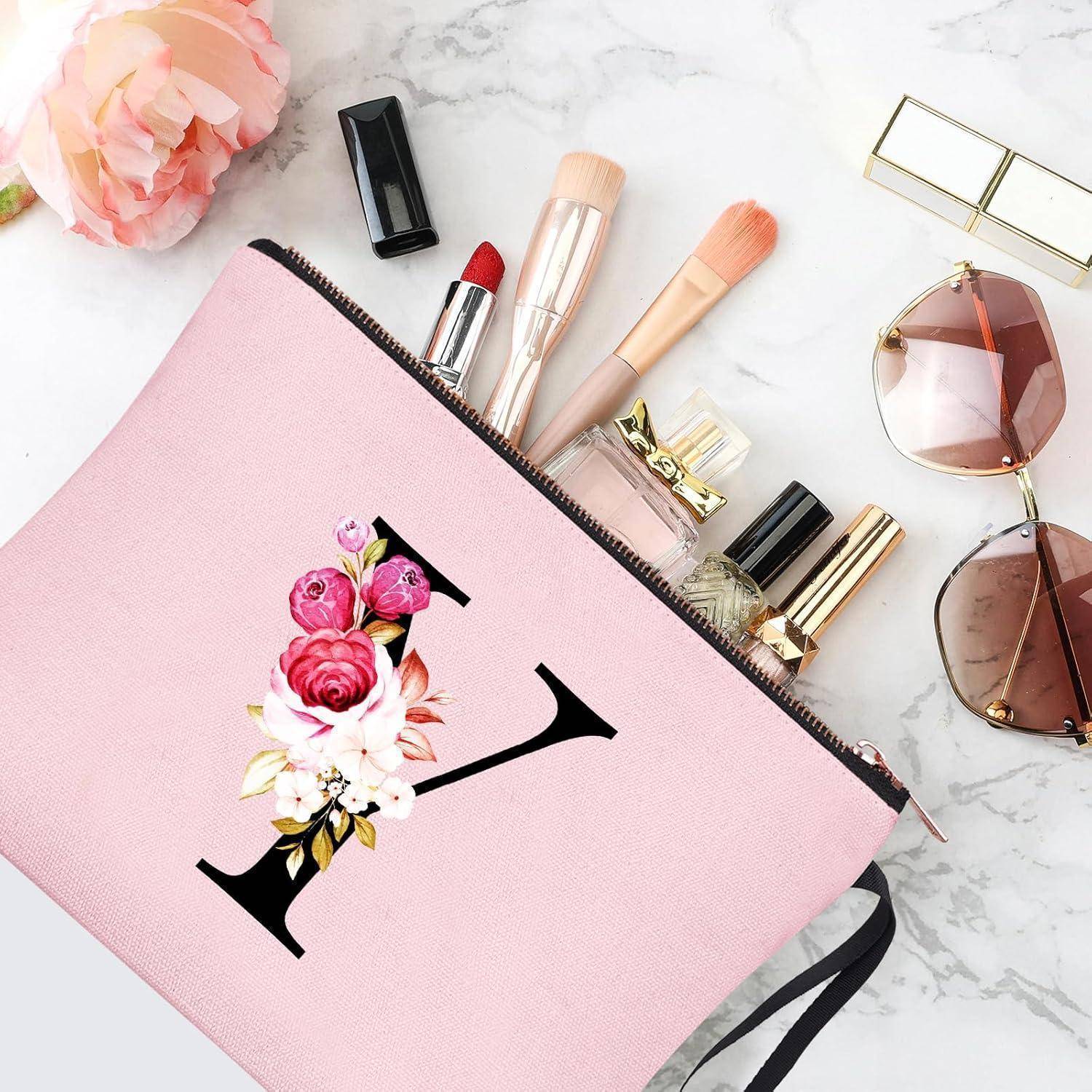 Eine rosa Schminkatasche mit Blumendekoration enthält Kosmetikutensilien, darunter Lippenstift, Pinsel und Nagellack. Eine Sonnenbrille und eine Blume liegen in der Nähe.