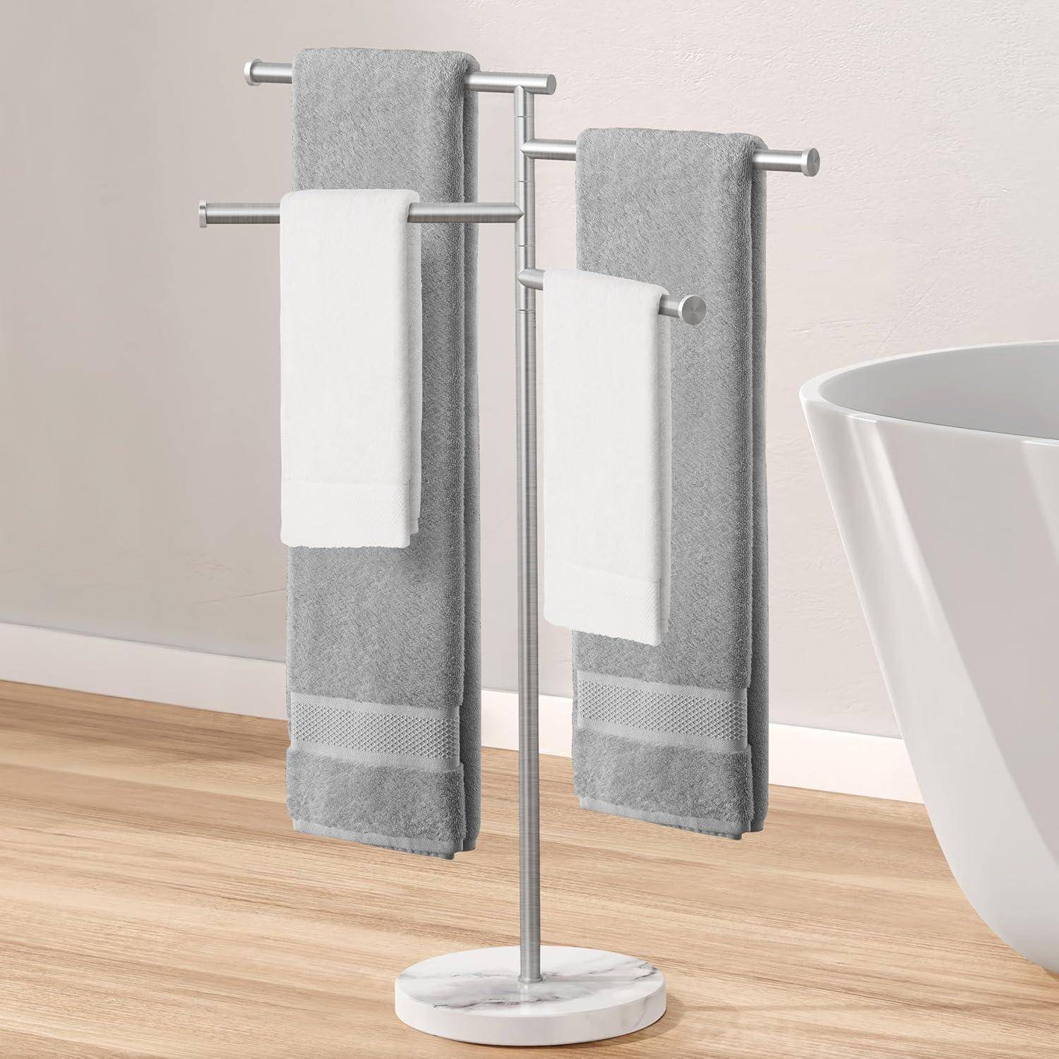 Ein Handtuchhalter aus Metall mit drei Stangen hält zwei graue Handtücher und zwei weiße Handtücher neben einer weißen Badewanne auf einem Holzboden.