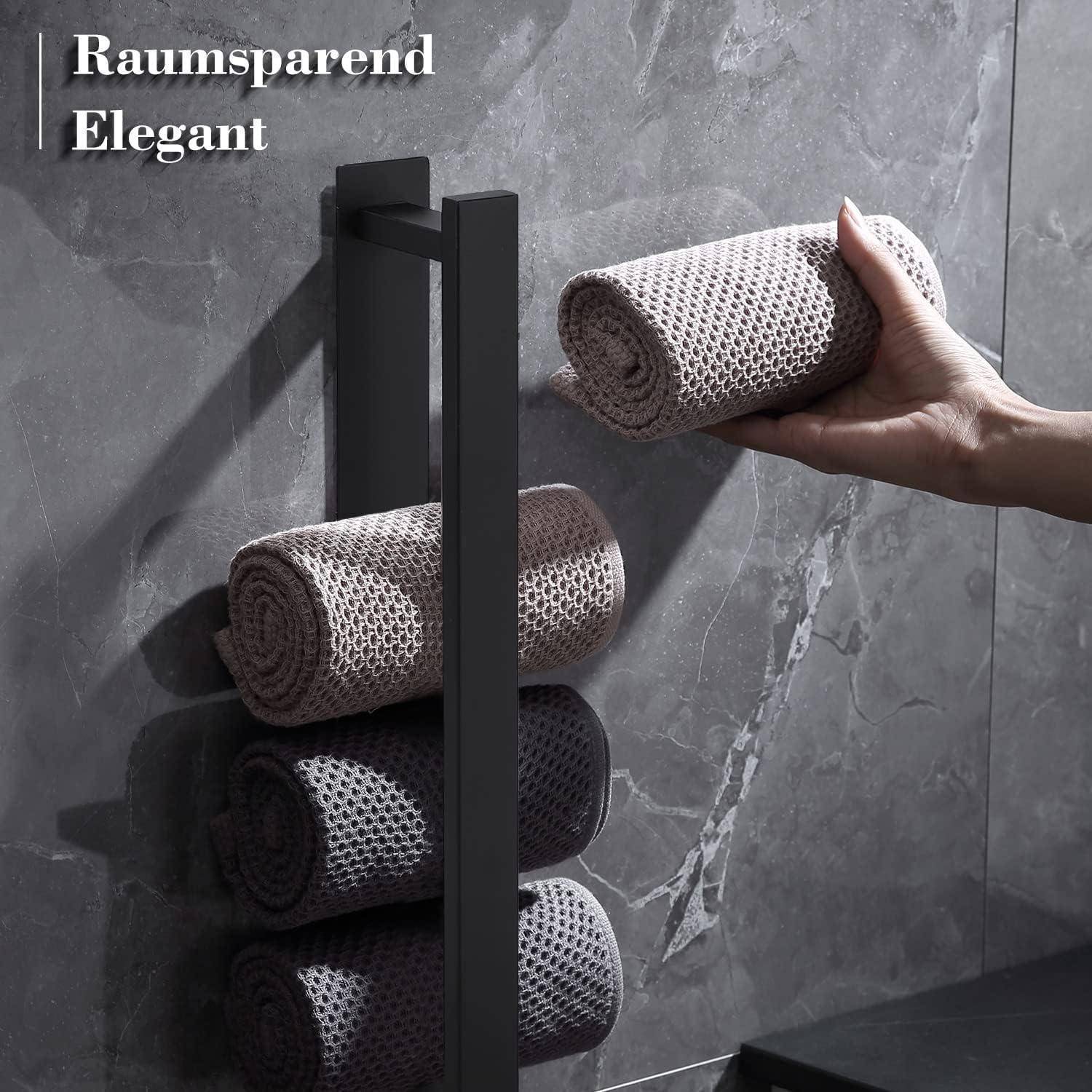 Eine Hand legt ein aufgerolltes Handtuch auf einen eleganten schwarzen Ständer vor einer grauen gefliesten Wand. Text liest sich: 'Platzsparend Elegant' in der oberen linken Ecke.