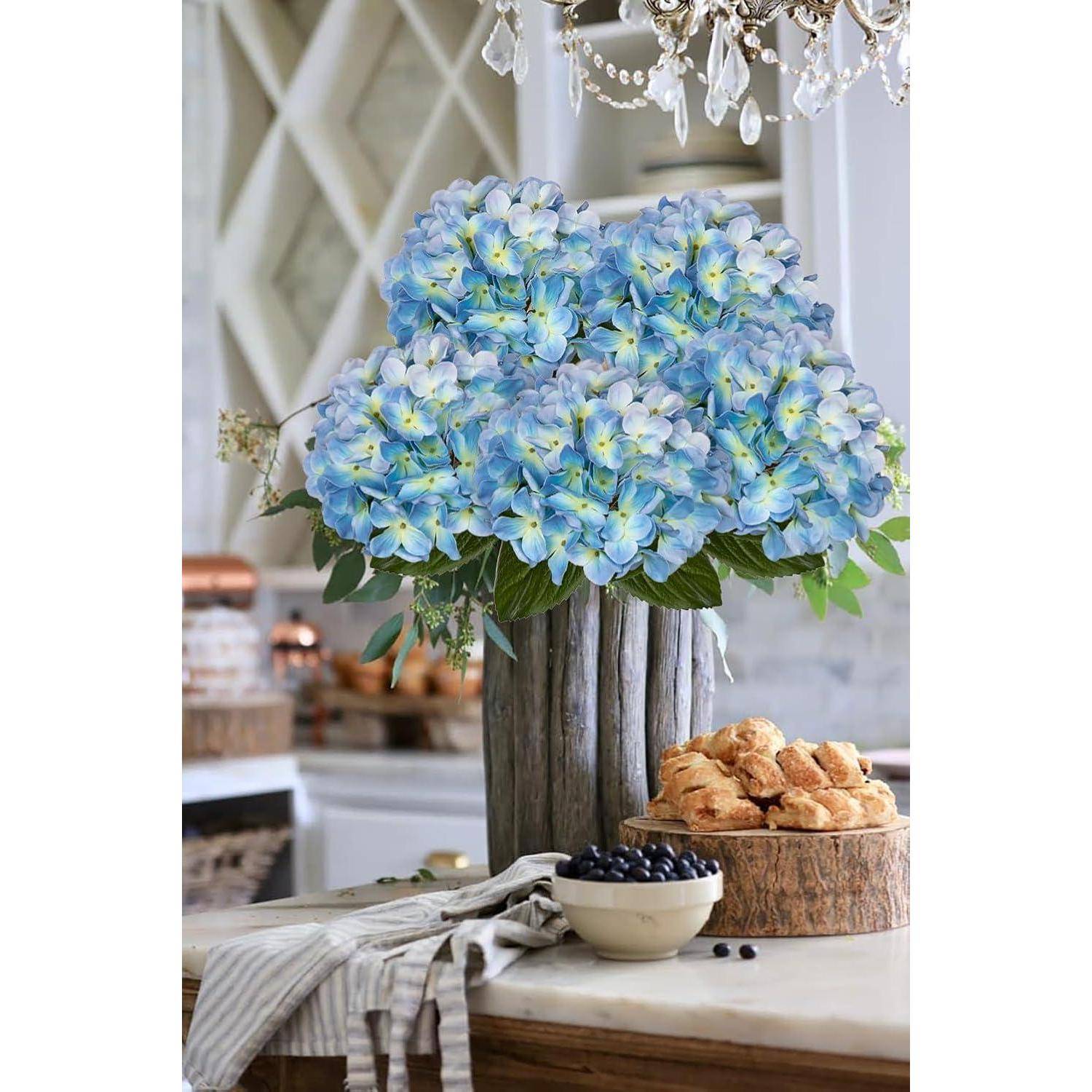 Eine rustikale Küchenarbeitsplatte mit einer großen Vase blauer Hortensien, einem Holztablett mit Croissants, einer Schüssel voller Blaubeeren und einem gestreiften Tuch.