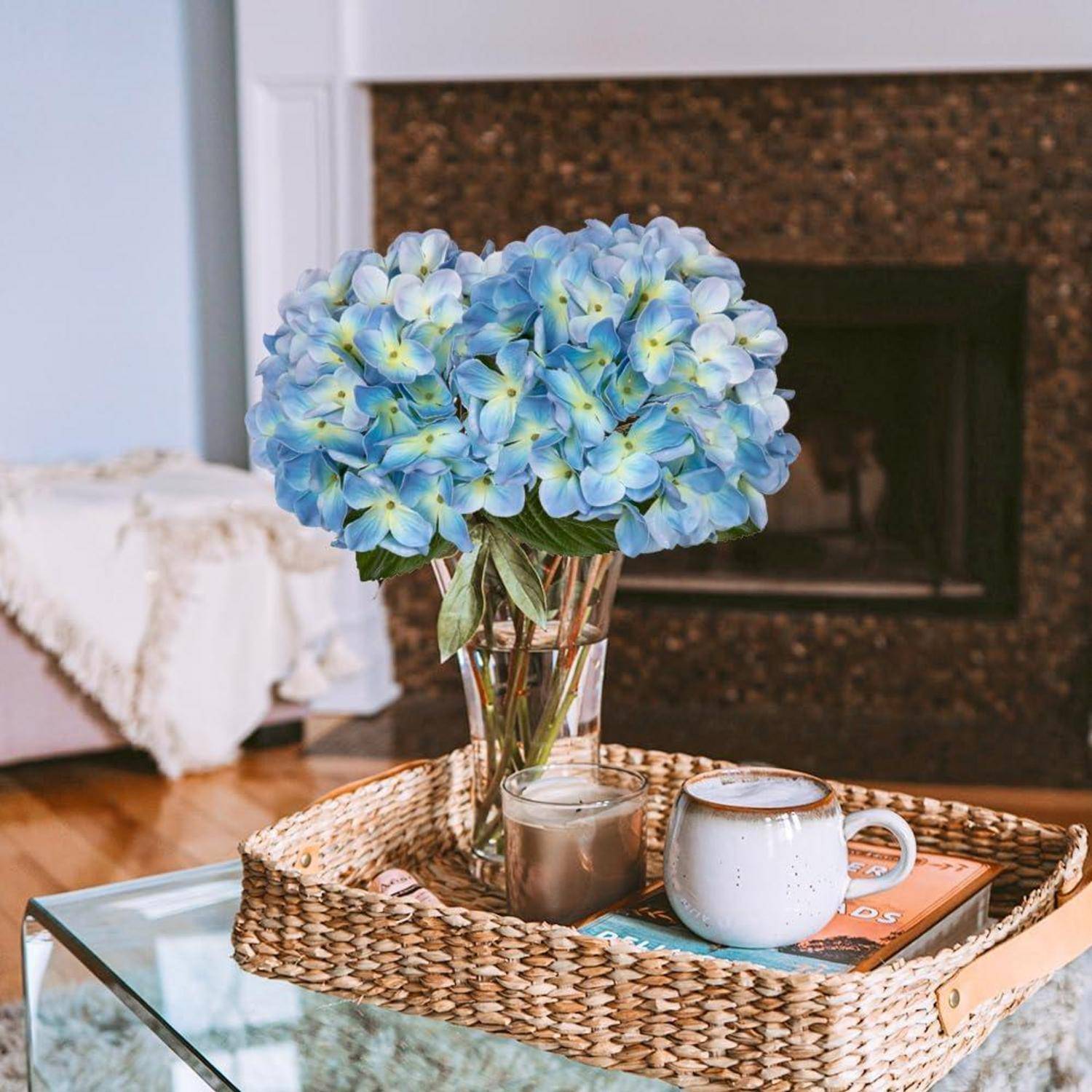 Eine Vase mit blauen Hortensien auf einem geflochtenen Tablett, daneben eine weiße Tasse und ein Buch, platziert auf einem Glastisch vor einem Kamin.
