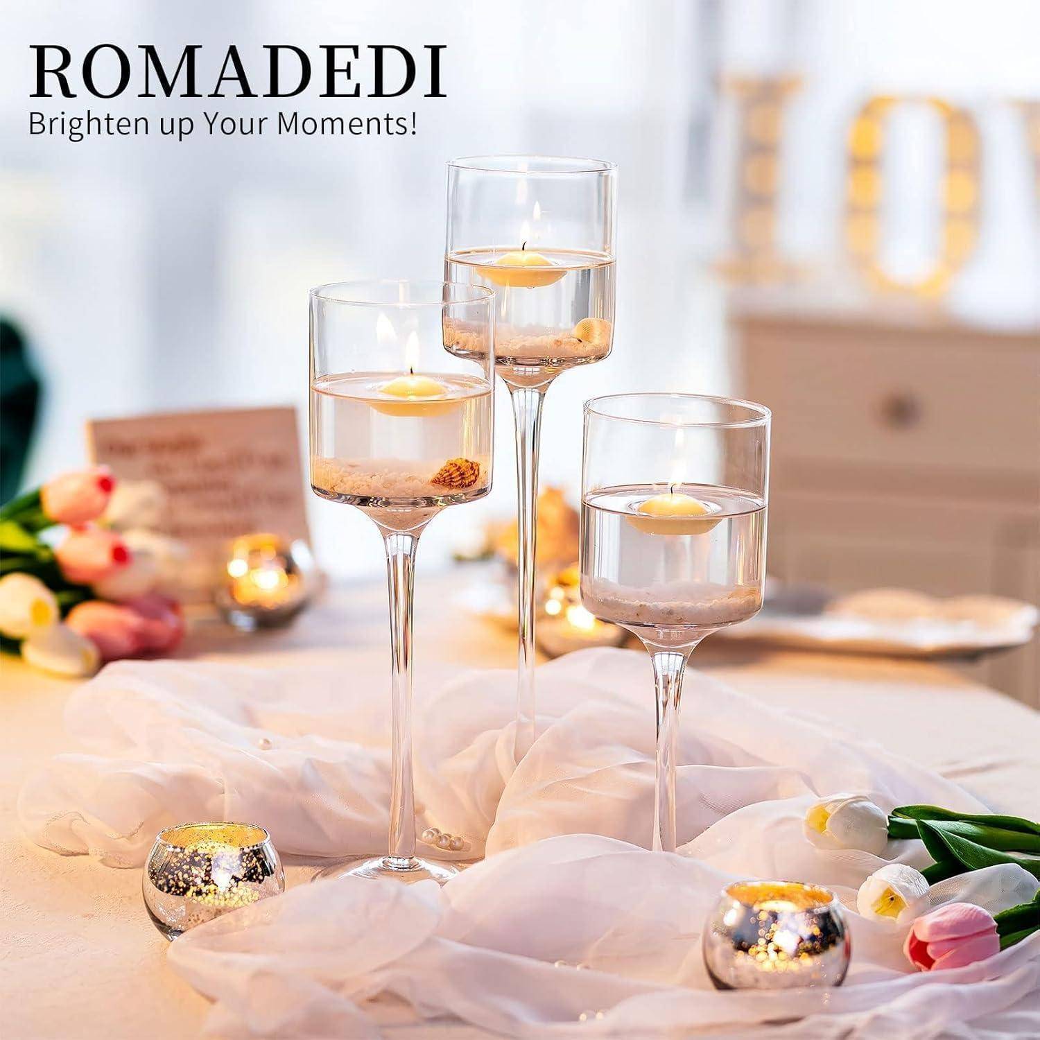 Drei elegante Kerzenhalter aus Glas mit schwimmenden Kerzen, umgeben von weichem rosa Stoff und kleinen Blumenarrangements, die eine romantische Atmosphäre schaffen.