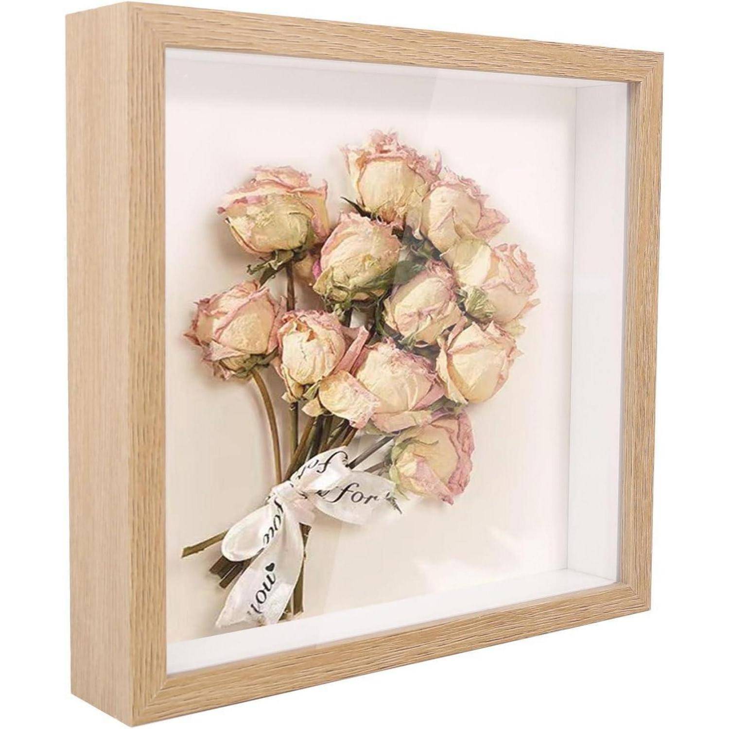 Ein gerahmtes Display aus getrockneten rosa Rosen, mit einem Band zusammengebunden, vor einem weißen Hintergrund. Der Holzrahmen verleiht dem Bild eine natürliche Note.