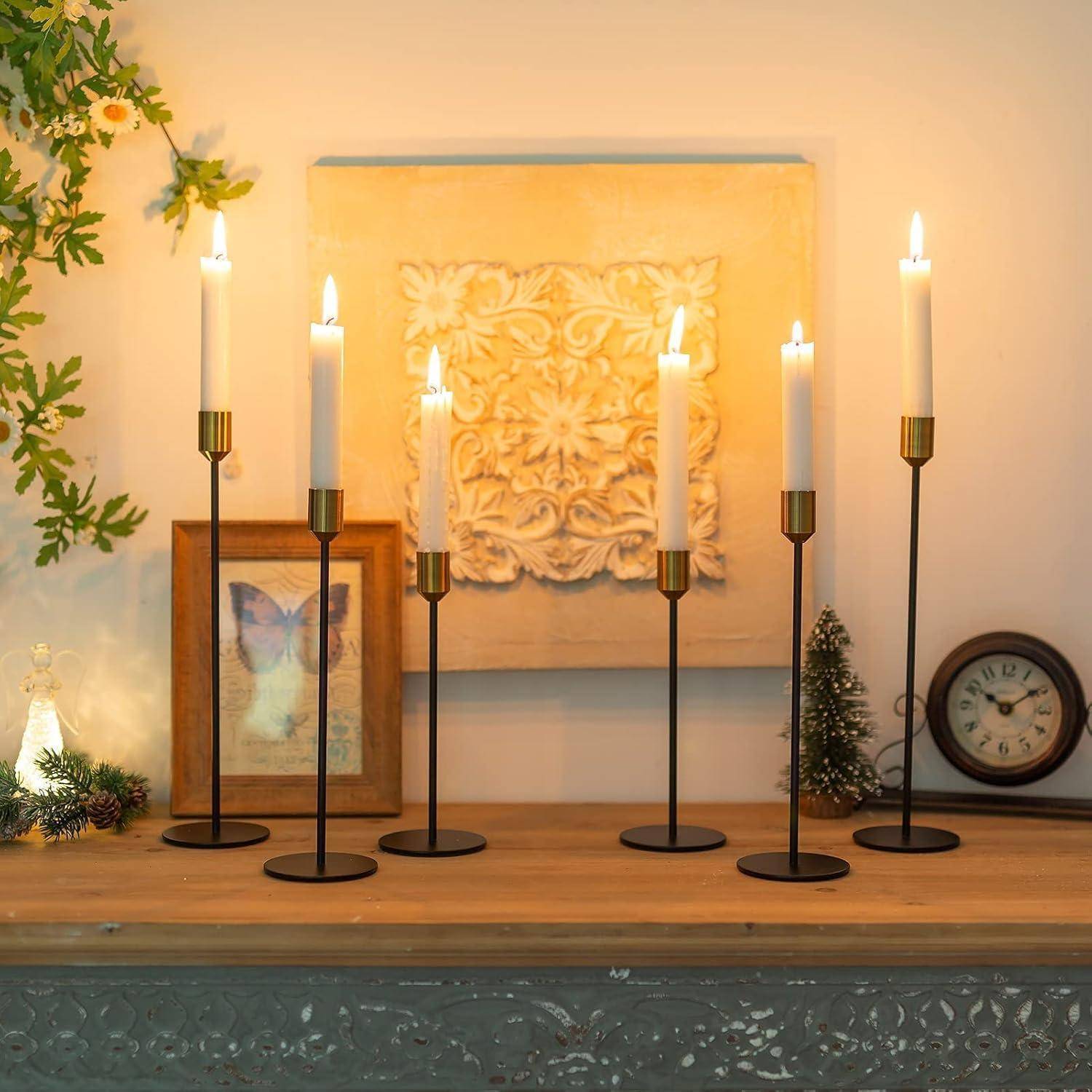 Eine Reihe von sechs brennenden Kerzen auf hohen schwarzen Halterungen auf dem Kaminsims, flankiert von kleinem Weihnachtsgrün, einer Uhr und gerahmter Kunst.