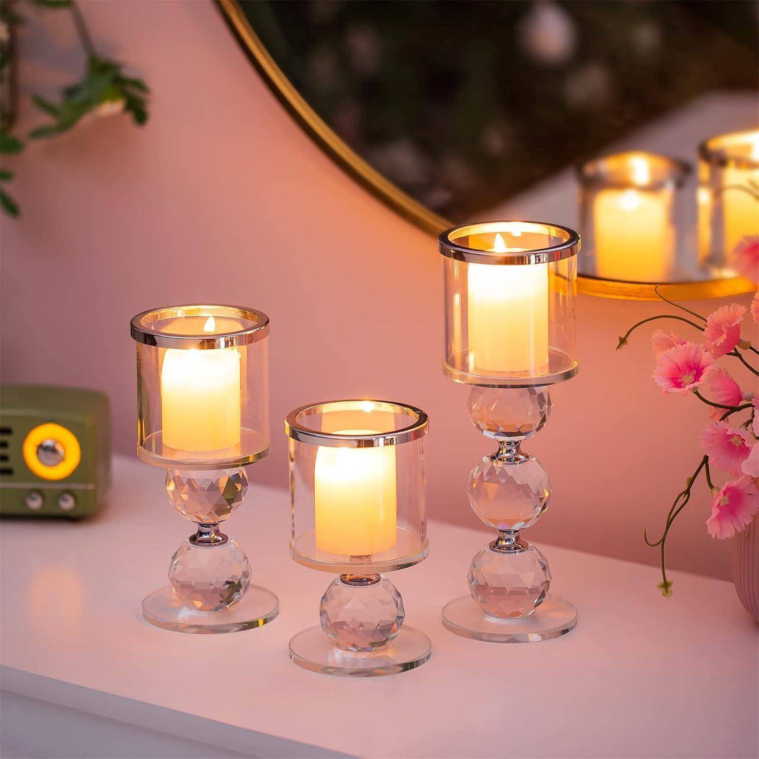 Drei elegante Glaskerzenhalter mit brennenden Kerzen stehen auf einer weißen Oberfläche und reflektieren warmes Licht. In der Nähe sorgen ein Vintage-Radio und rosa Blumen für eine gemütliche Atmosphäre.