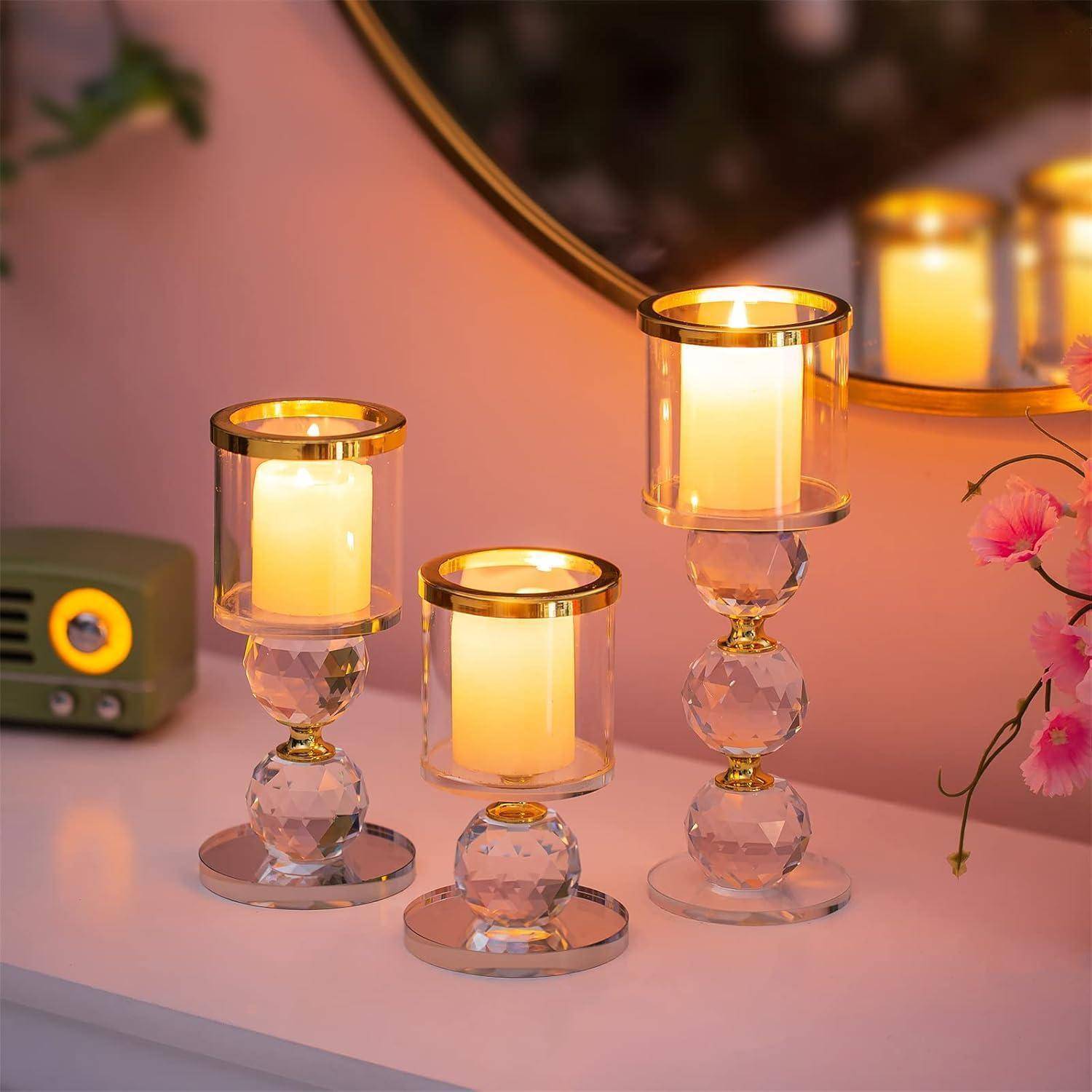 Drei dekorative Glaskerzenhalter, in denen jeweils eine brennende Kerze steht, sind auf einer weißen Oberfläche neben einem kleinen grünen Radio und rosa Blumen platziert.