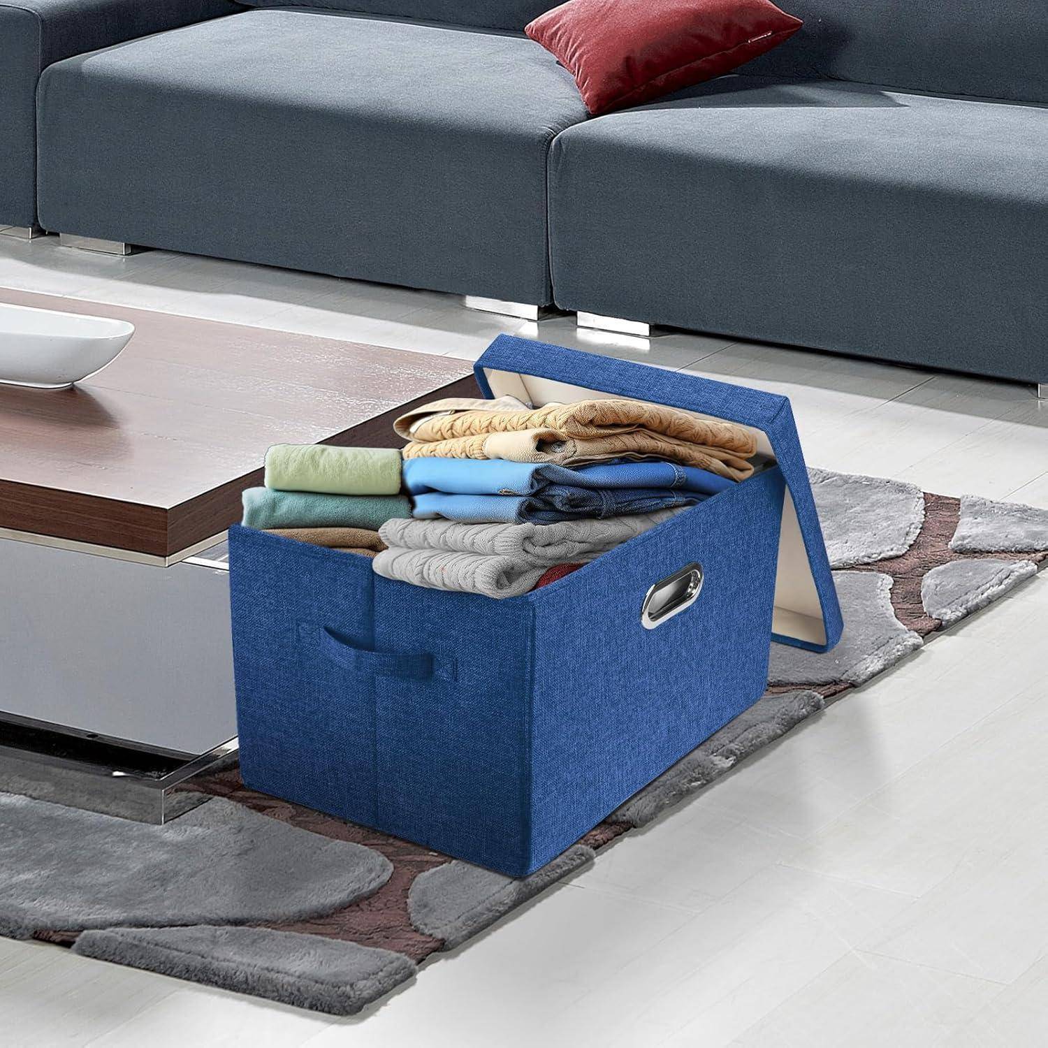 Eine blaue Aufbewahrungsbox, gefüllt mit zusammengelegten Kleidungsstücken, steht auf dem Boden neben einem modernen Couchtisch und einem grauen Ecksofa in einem Wohnzimmer.