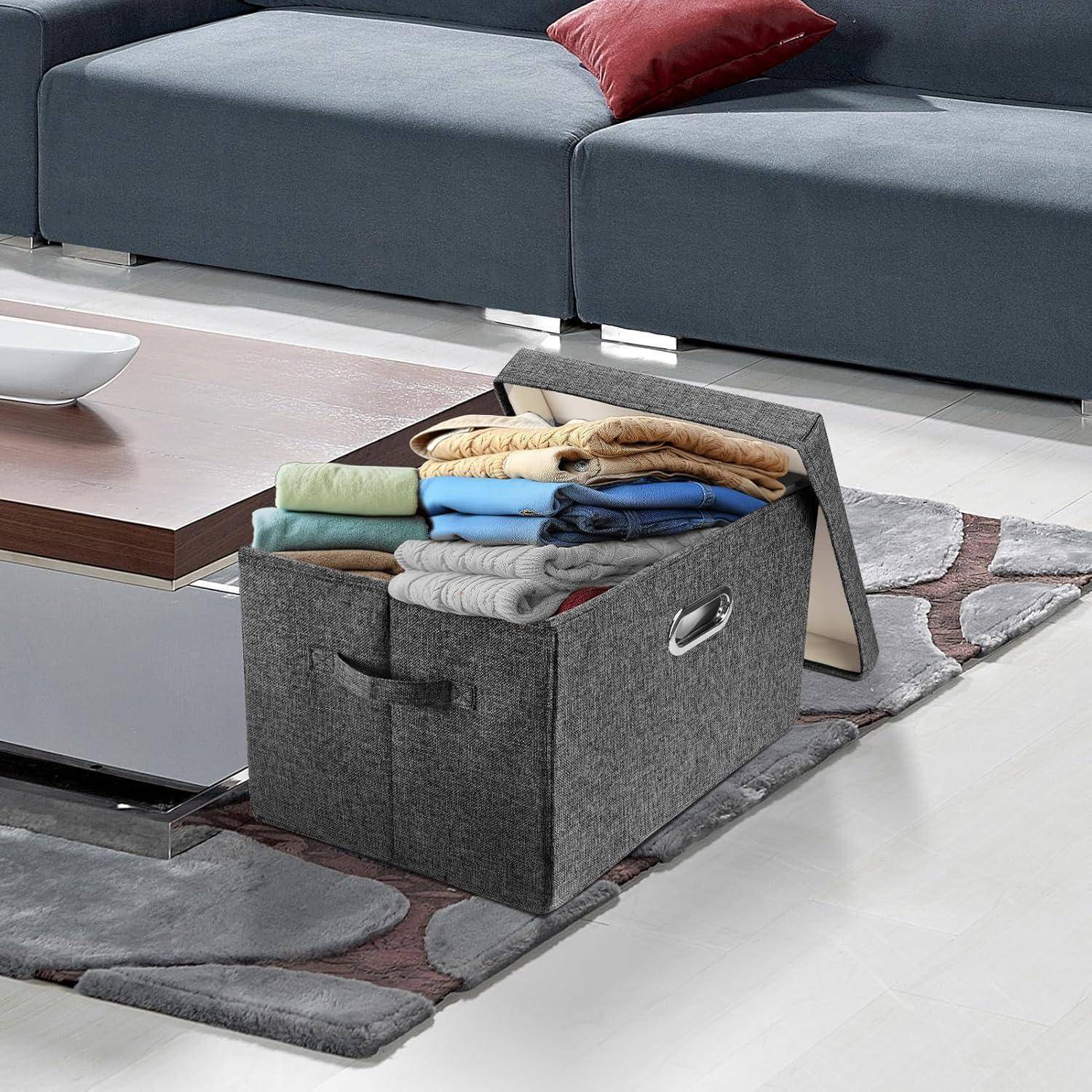 Eine graue Aufbewahrungsbox, gefüllt mit ordentlich zusammengelegten Kleidungsstücken, steht auf einem modernen, hellen Boden neben einem dunkelblauen Sofa mit einem roten Kissen.