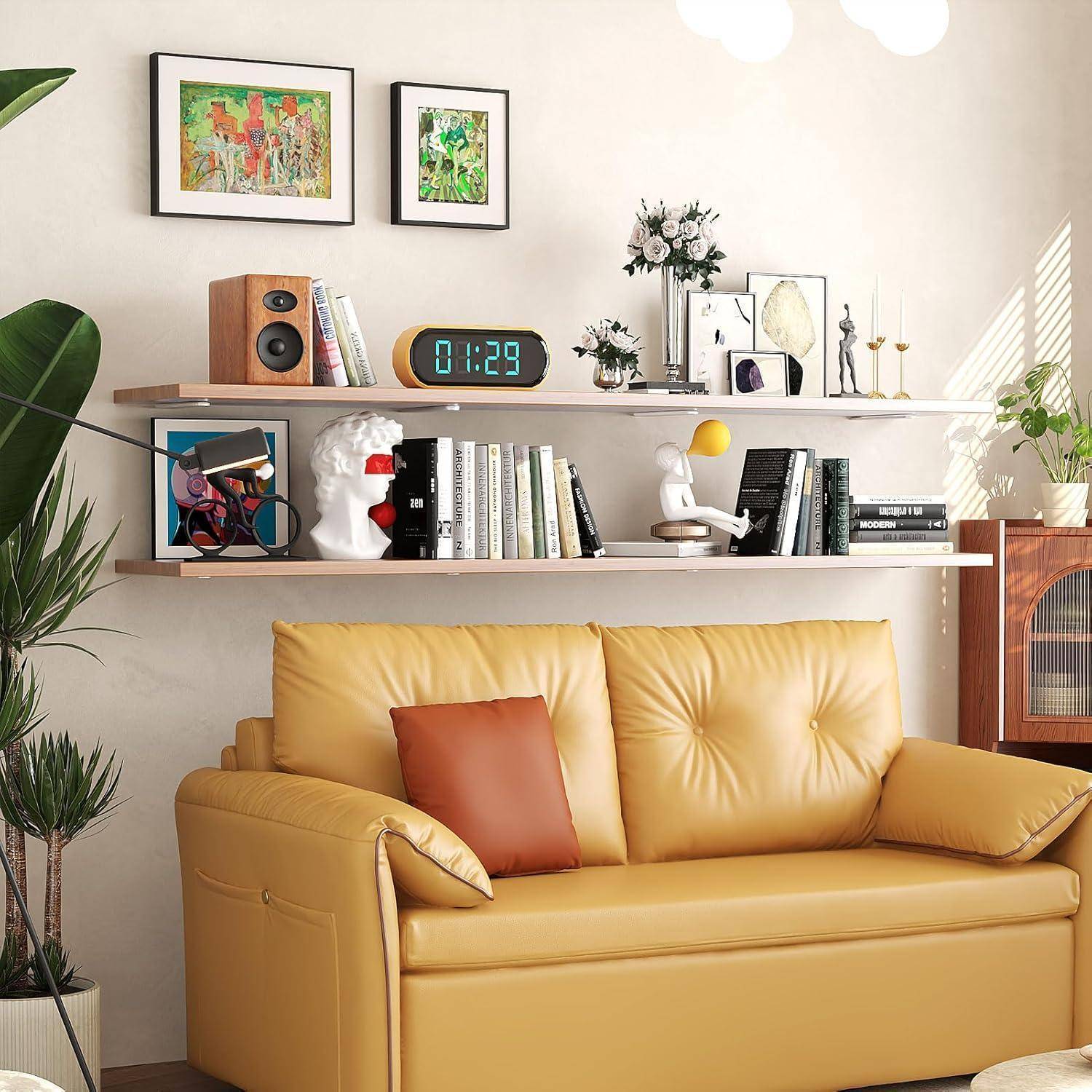 Ein gemütliches Wohnzimmer zeigt eine beige Ledercouch mit orangefarbenen Kissen. Regale darüber präsentieren Bücher, Pflanzen und Dekorationsgegenstände. Sonnenlicht strömt herein.