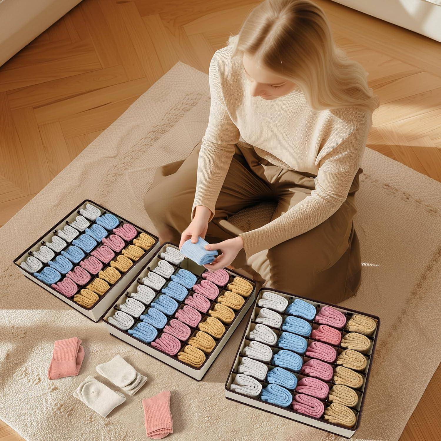 Eine Person sitzt auf dem Boden und sortiert pastellfarbene Socken in drei Kisten, während weitere Socken auf einem beigen Teppich verstreut sind.