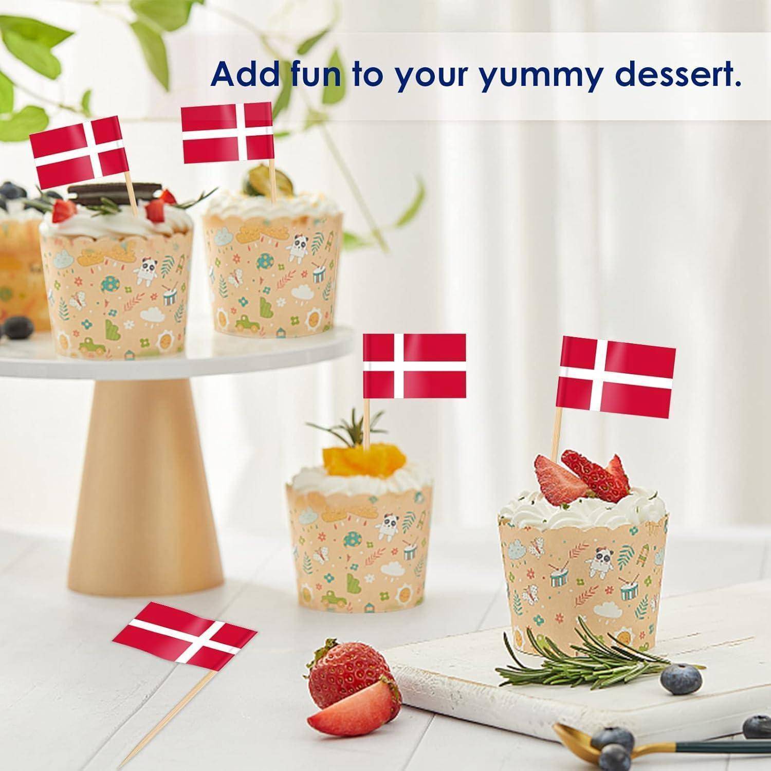 Drei Cupcakes mit dänischen Flaggen oben auf einem Tisch, umgeben von Früchten. Text lautet: 