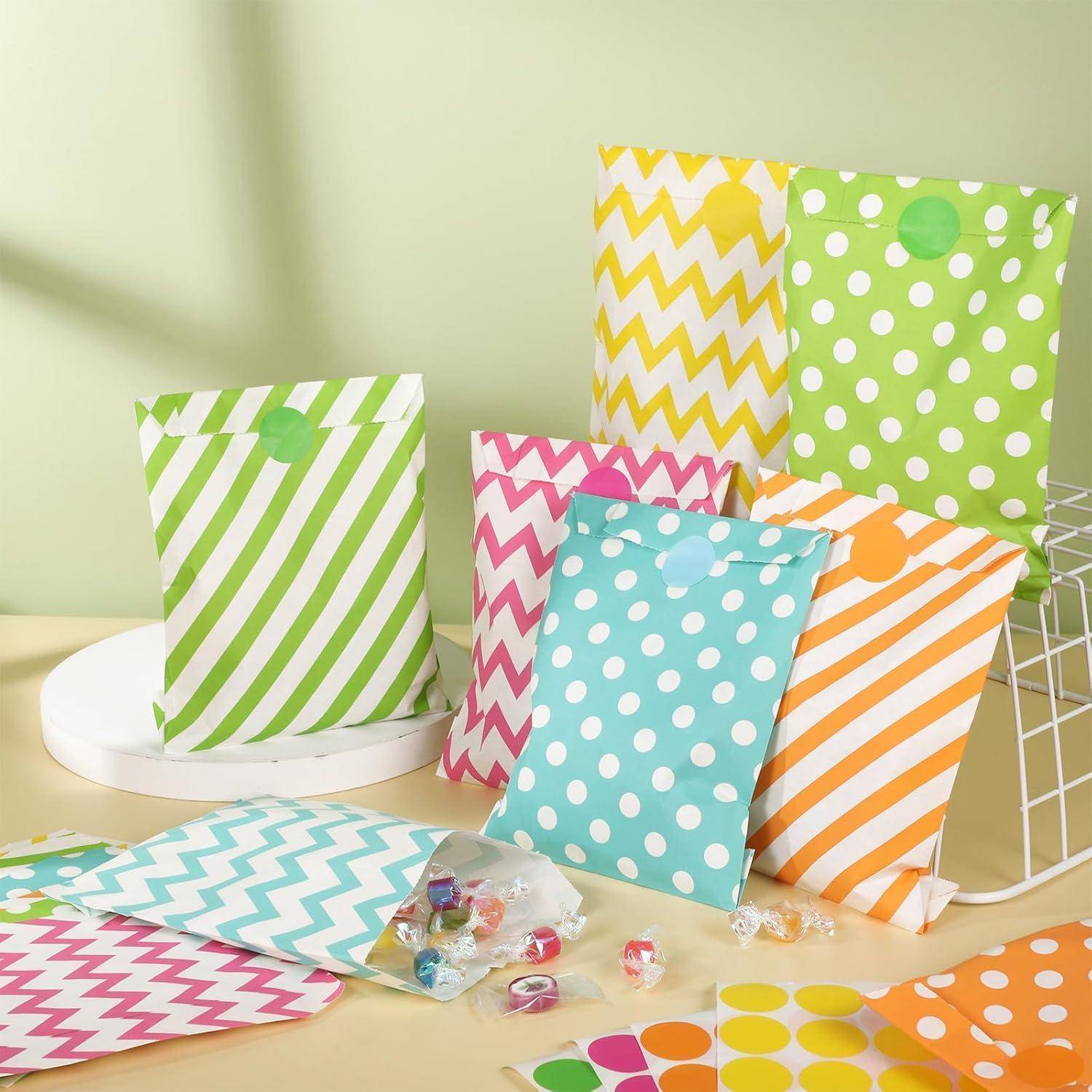 Bunte gestreifte und gepunktete Geschenktüten in Grün, Pink, Blau, Orange und Gelb auf einem Tisch, mit kleinen Süßigkeiten und Stickern, die in der Nähe verstreut sind.